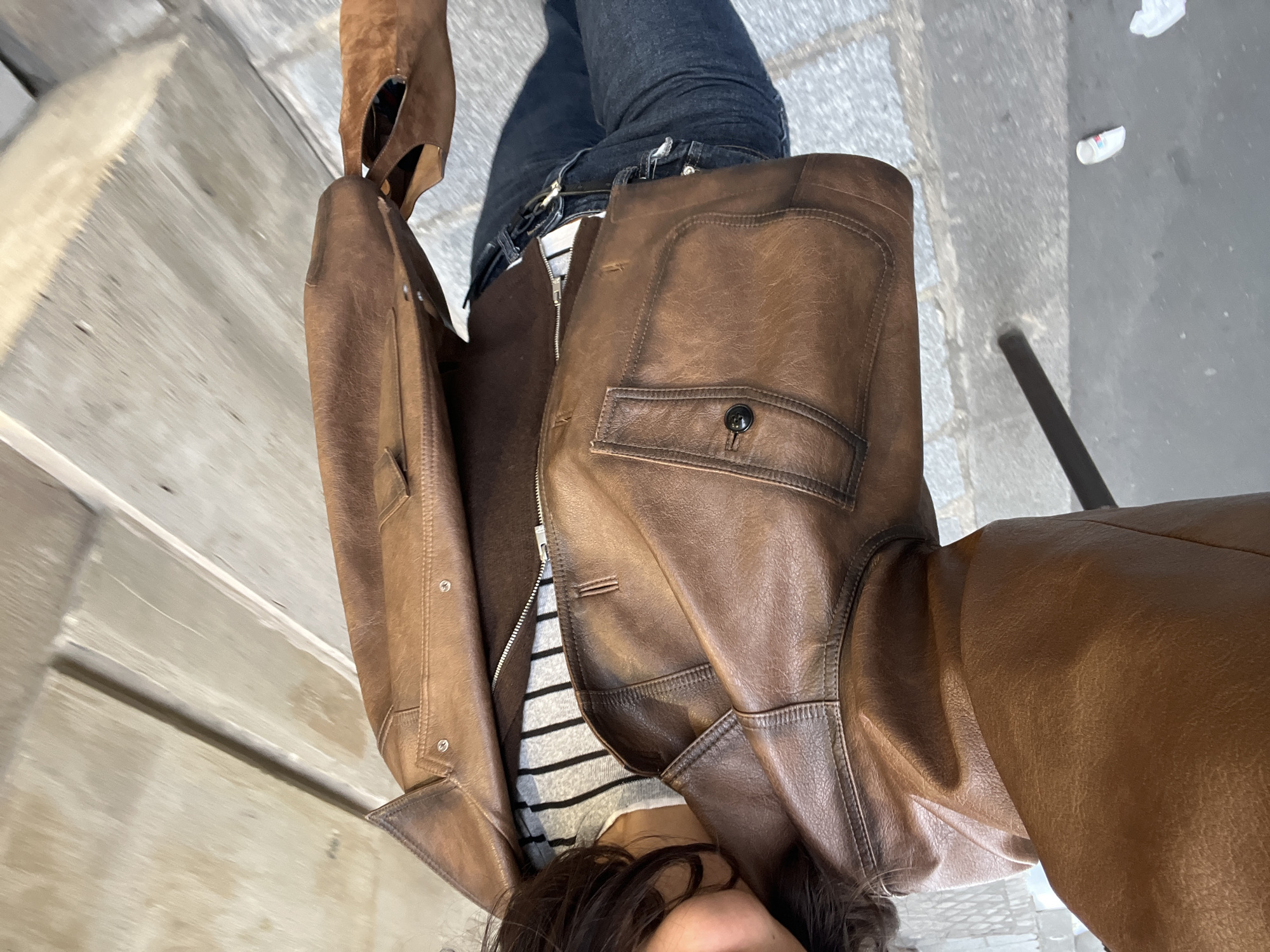 An individual is depicted in a waist-up selfie on a paved street in Paris, France. The person has short, dark, wavy hair and visible lips with a slight upturn, suggesting a subtle smile. They are dressed in a layered outfit, consisting of a brown leather jacket, a brown zippered garment underneath, and a white and grey horizontally striped shirt. The leather jacket features a spread collar, two buttoned flap pockets on the chest (the left one visible with a dark button), two lower patch pockets (the left one visible), and metal studs along the seam of the right upper arm. A dark belt with a light-colored buckle is visible above dark wash jeans. The person's right hand holds a light brown, suede-like tote bag with cut-out handles, revealing a striped interior lining. The ground is composed of grey stone pavers. A dark vertical pole is visible on the left, near which a small white cup with colored stripes and a piece of crumpled white paper are discarded on the pavement. In the background to the right, a light-colored building facade or door, possibly wood or painted plaster, shows distinct horizontal and vertical paneling. Natural light illuminates the scene, casting soft shadows.