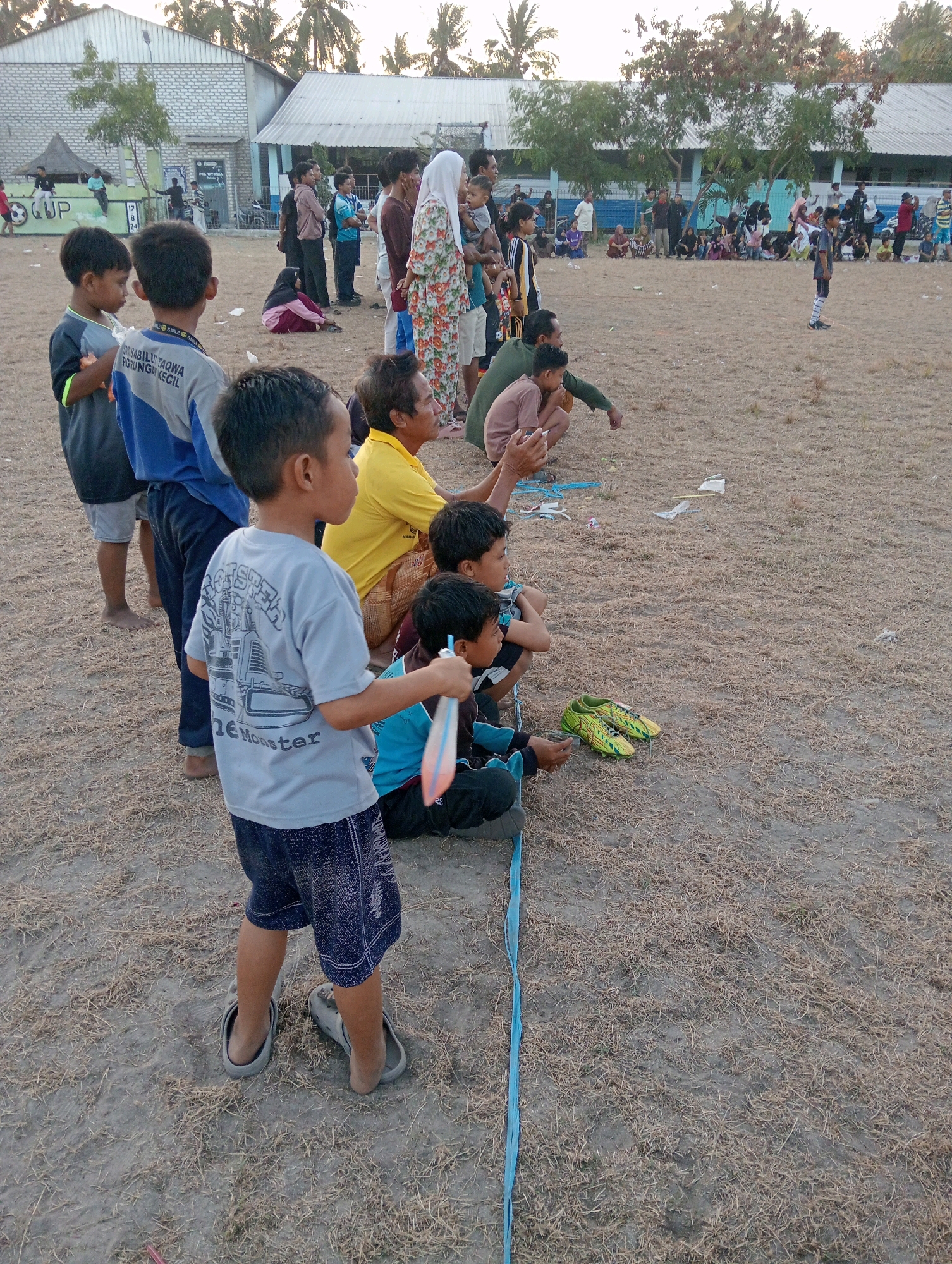 The image depicts an outdoor scene in Pagerungan Kecil, Indonesia, likely during an event. In the foreground, several boys are seated or standing on a dry, dirt field covered with sparse, dead grass. One boy in a light blue t-shirt with a graphic of a bulldozer and the text "Monster" is prominently featured. He is wearing dark blue shorts and grey clogs. Another boy in a blue and grey shirt is standing next to him. Further back, more children and adults are visible, some sitting on the ground and others standing. In the middle ground, a group of adults and children are gathered, with some individuals wearing traditional Indonesian clothing. In the background, there are buildings with white and blue facades, possibly school buildings or community centers, with palm trees visible overhead. The overall atmosphere appears to be casual, with people observing an activity or event taking place on the field. There are scattered pieces of debris, including paper and plastic, on the ground. A bright blue ribbon or cord runs diagonally across the foreground of the image.