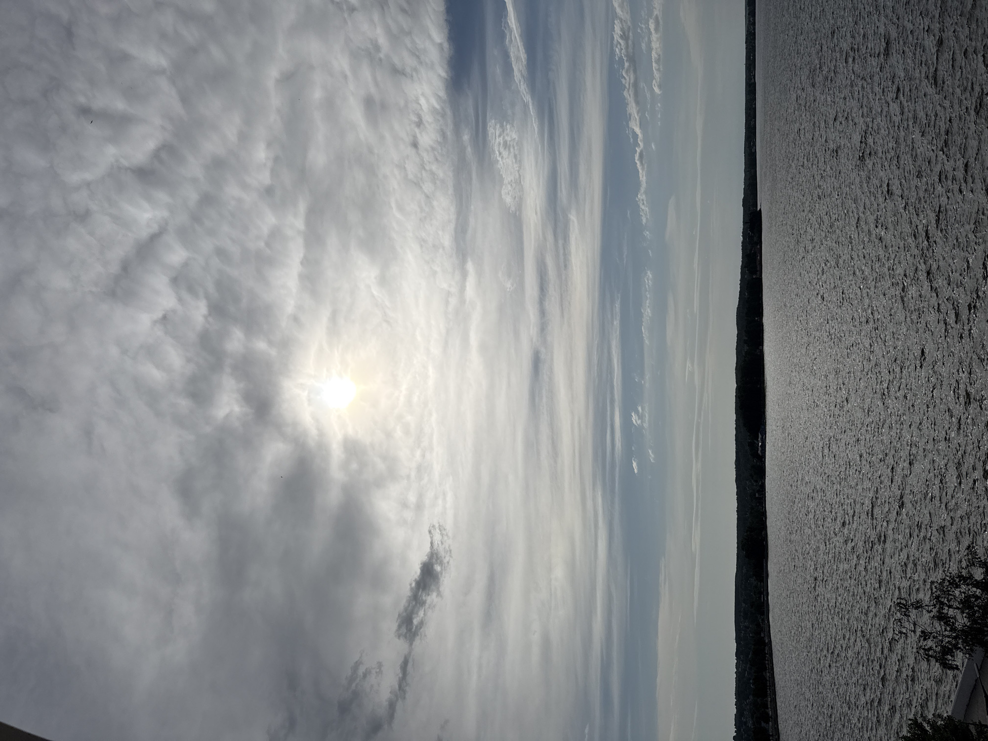 The image shows a body of water, likely the Votkinsk Reservoir, under a cloudy sky with the sun visible through the clouds. A strip of land, potentially an island or shoreline, is visible between the water and the sky. The location is likely near Votkinsk, Russia, given the reservoir's location. There are no people visible in the frame. The water's surface is rippled, reflecting the sunlight. The clouds vary in texture and density. Some foliage is visible in the lower right corner, suggesting a nearby tree or bush. The sky occupies a significant portion of the image.