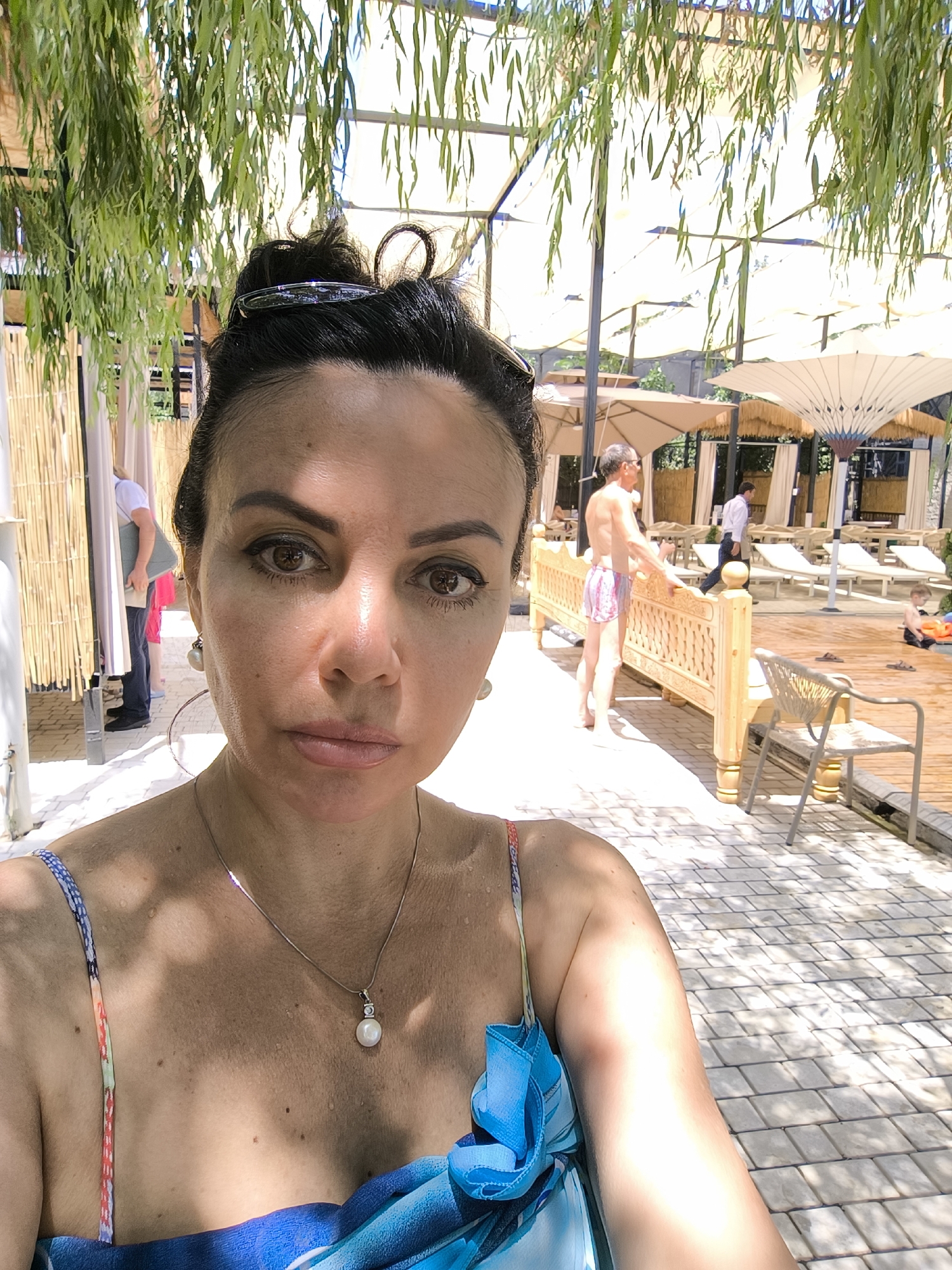 A woman takes a selfie outdoors in what appears to be a resort or spa area. She is facing the camera, and her upper body is visible, wearing a blue and colorful patterned swimsuit. She has dark hair pulled up, sunglasses on top of her head, and is wearing a pearl pendant necklace and large hoop earrings. Behind her, a man in pink and white patterned shorts stands near a wooden bench. Other people, lounge chairs, and umbrellas are visible in the background, suggesting a recreational setting. The ground is paved with light-colored tiles. The location is identified as the Vahdat city, Tajikistan.