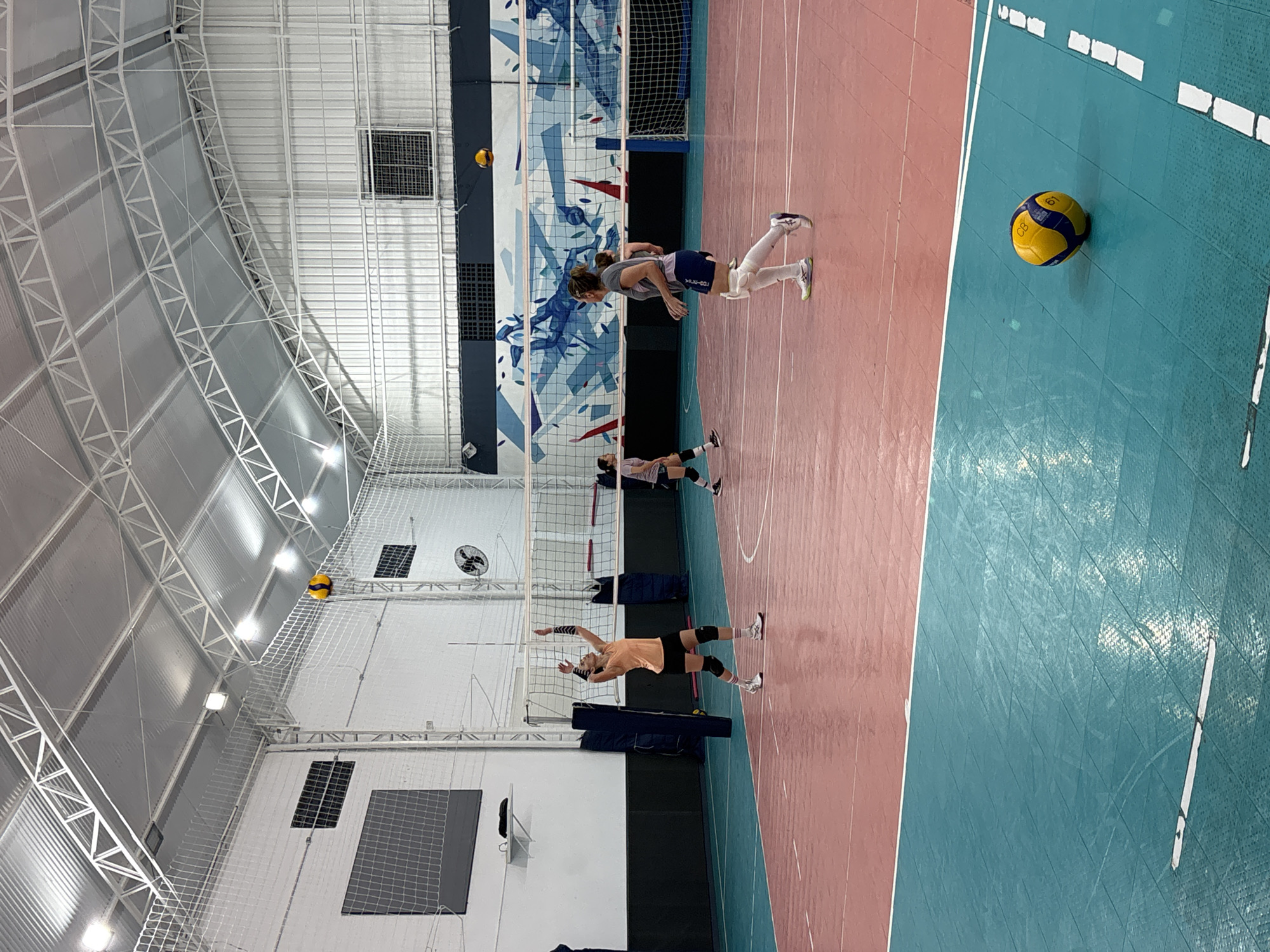 The image shows an indoor volleyball court, likely a training or practice space, with a volleyball in the foreground on the blue section of the court. The court is lined with red and blue sections, separated by a white line. Three people are visible in the midground, positioned on different sections of the court; one appears to be in the act of hitting a volleyball, while the others are in a ready stance. A volleyball net stretches across the middle of the court, and artwork or a design is visible behind the net. The background shows a large, arched building structure with white support beams, lighting fixtures, and ventilation elements. The setting is strongly suggested to be in São Paulo, Brazil, given the context and court design that are often used for volley ball courts in this city.