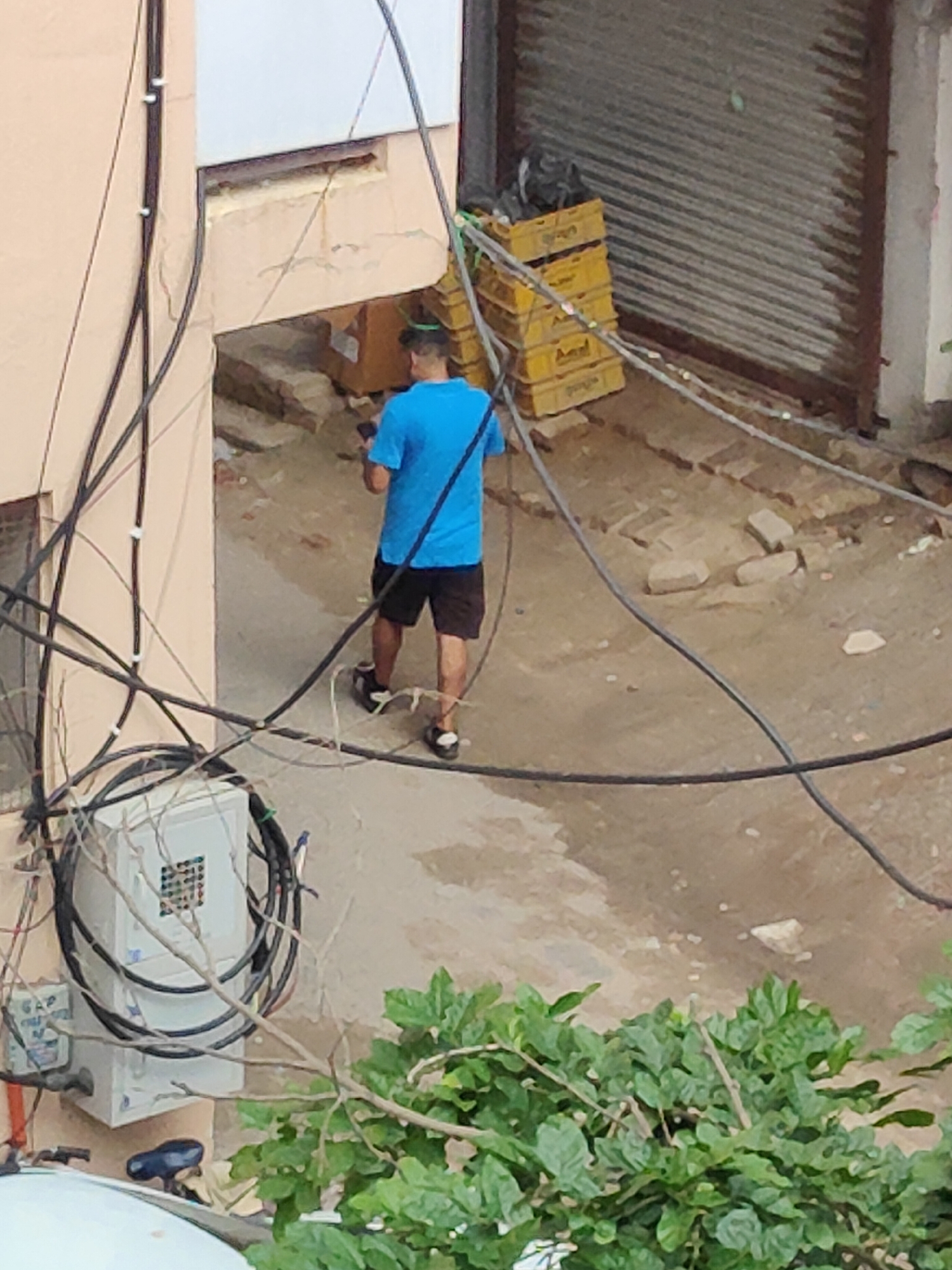 The image depicts an outdoor scene in Gurgaon, India, characterized by a cluttered urban environment. In the foreground, a person is seen walking away from the camera, wearing a blue t-shirt and black shorts. The person is holding a dark-colored object, possibly a phone, in their right hand. The ground is uneven and appears to be a mix of dirt and some debris.
The backdrop features a building with a light-colored facade and an open doorway. Adjacent to this, a metal roll-up shutter is visible, with stacks of yellow plastic crates to its left. Numerous black electrical wires and cables are strung across the scene, some attached to the building's exterior and others suspended in the air. A white electrical box is mounted on the wall, with several wires coiled around it. In the lower right corner, lush green foliage is present, partially obscuring the scene. The lighting suggests it is daytime.