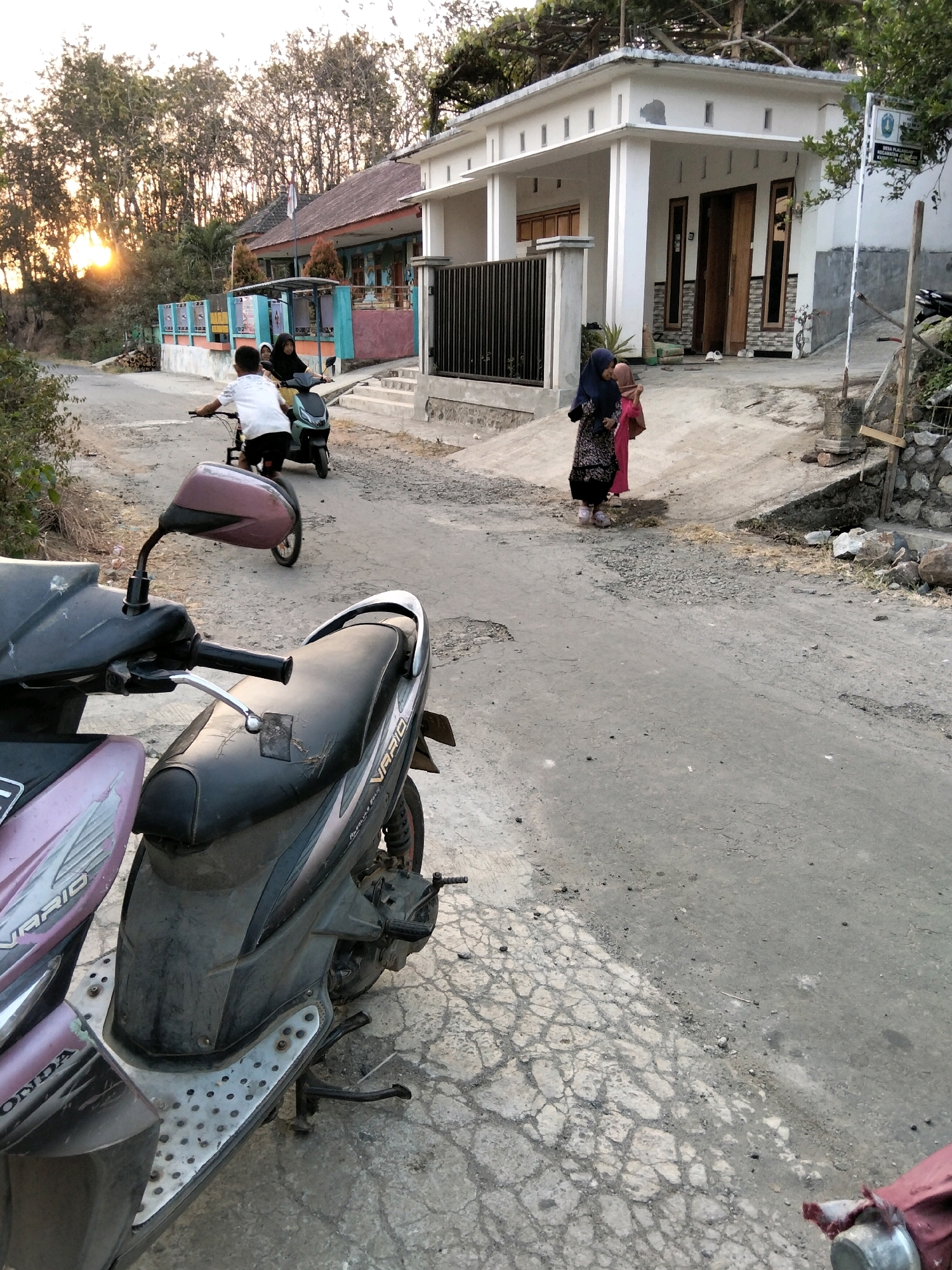 In the city of Jenangan, Indonesia, a rural road scene unfolds at dusk. In the foreground, two parked motorcycles, one pink and one dark, dominate the perspective, their seats worn and dusty. The road surface is cracked and uneven, with scattered gravel and debris. In the middle ground, three individuals are present. A young boy on a bicycle, wearing a white shirt and dark shorts, cycles away from the viewer. Behind him, on a dark scooter, a young girl in a headscarf and a woman in a patterned dress and headscarf are seated. They are facing the viewer, and the woman appears to be speaking to the girl. The background features a modest, white, single-story building with a tiled roof, possibly a home or small business, and a fence. To the left, trees with dense foliage line the road, and the setting sun casts a warm glow on the horizon. The overall impression is of a quiet, everyday moment in a village setting.
