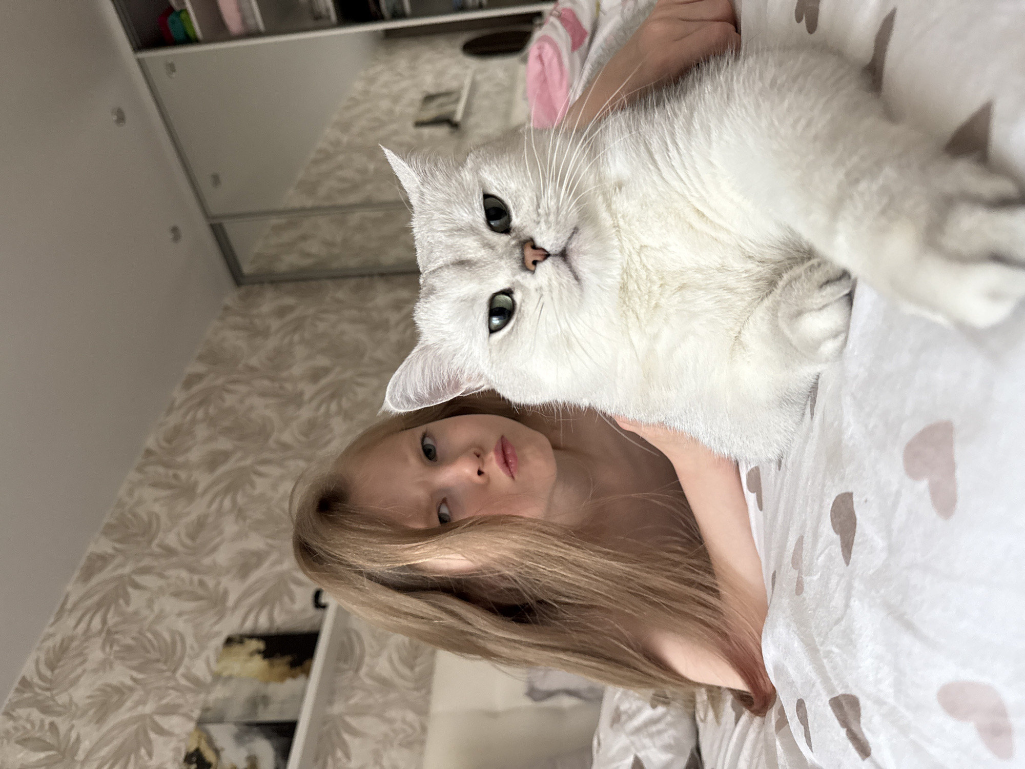 A blonde-haired girl is lying on a bed with a white cat. The cat is white with green eyes and is lying on the girl's chest. The bed has a white comforter with gray heart patterns. The background includes a wall with leaf-patterned wallpaper and a white closet. The location cannot be determined from the image, although requested.