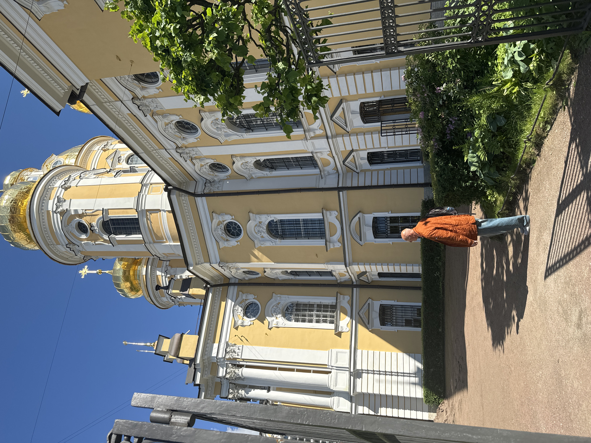 A man in an orange jacket and blue jeans stands facing a yellow church with golden domes against a clear blue sky. The church features ornate white trim and tall windows. A black metal fence is visible on the left and right sides of the image, partially obscuring the building and surrounding greenery. The ground is covered with reddish-brown gravel, and shadows from trees and structures are cast across it. The scene appears to be in a courtyard or garden area adjacent to the church, which is likely in Saint Petersburg, Russia, given the architectural style and golden domes characteristic of Russian Orthodox churches.