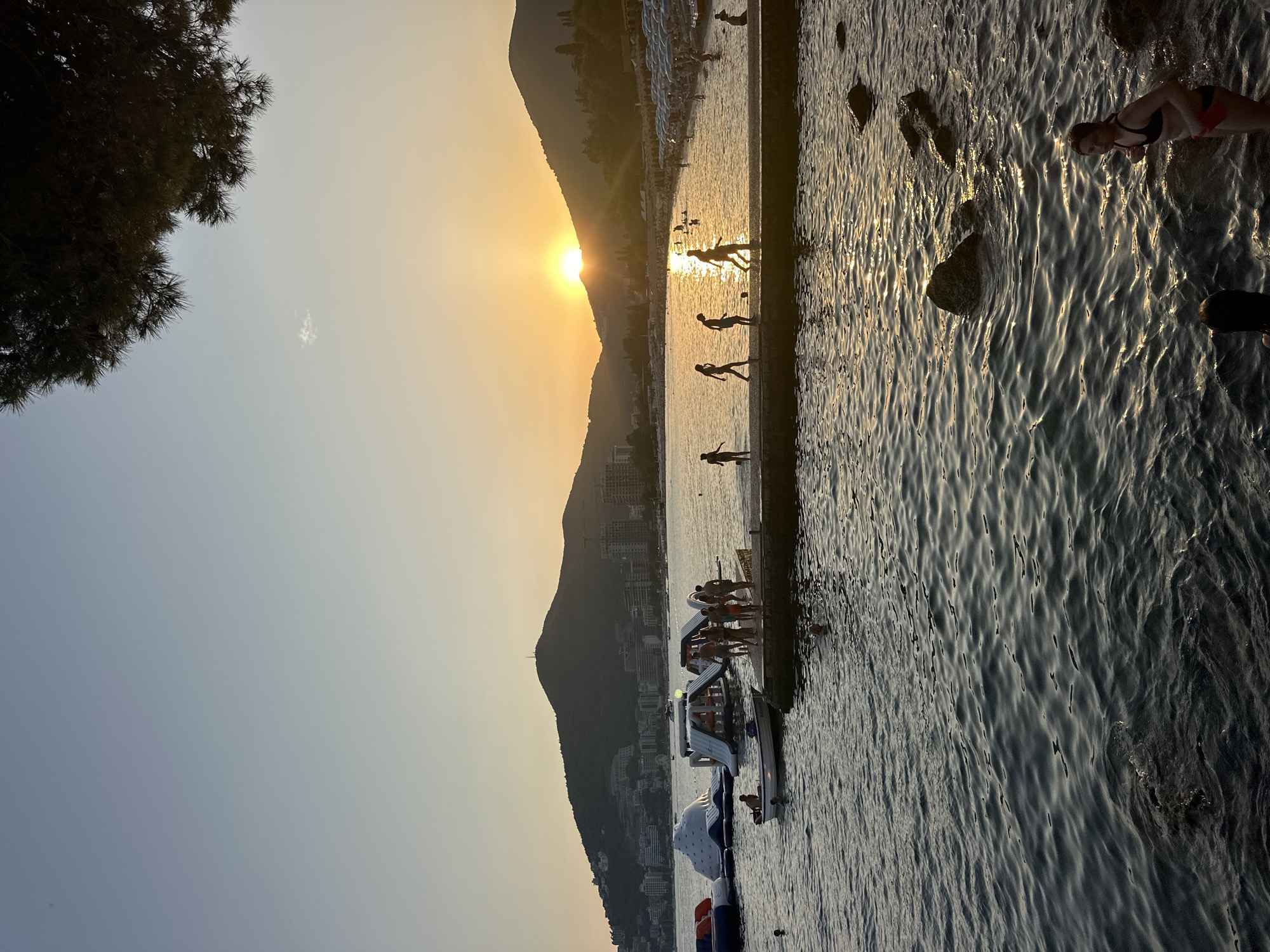 The image depicts a sunset scene at Mogren Beach in Budva, Montenegro. The sun is setting behind a mountain range, casting a warm glow over the water and the landscape. In the foreground, the dark, rippling surface of the Adriatic Sea is visible, with several large rocks emerging from the water on the right. A young woman in a black bikini and red bottoms stands in the water on the right, looking towards the camera.
In the mid-ground, a concrete pier extends into the water, with several people walking on it. Some are silhouetted against the setting sun, and a few appear to be playing with a ball. To the left of the pier, several inflatable water structures, possibly part of a water park, are floating in the sea. A small white boat is tied to one of these structures.
The background shows a coastline lined with buildings and more mountains. The sky transitions from a pale blue at the top to a warm yellow and orange near the horizon where the sun is setting. The overall atmosphere is serene and captures a typical summer evening at a popular beach destination.