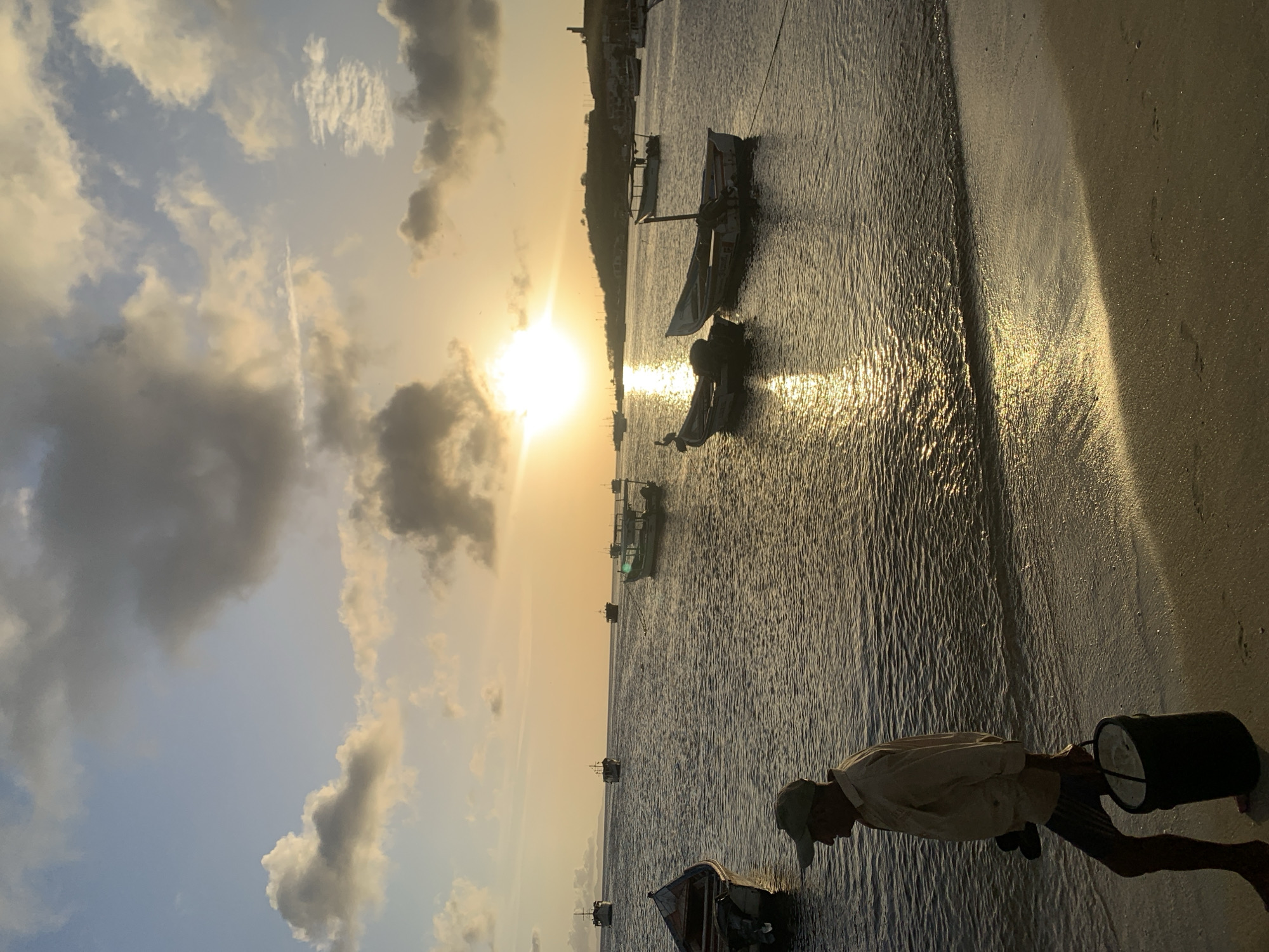 The image depicts a serene sunset scene at the coast of Juan Griego, Venezuela. A man, wearing a light-colored shirt, shorts, and a baseball cap, is walking along the sandy shore. He is carrying a dark bucket in his right hand and a pair of sandals in his left. The man is walking from left to right, towards the right edge of the frame. The sun is low in the sky, casting a golden glow on the water and the wet sand. The ocean is calm, with small waves lapping at the shore. Several fishing boats are anchored in the water, some closer to the shore and others further out. In the background, a hilly coastline with scattered buildings is visible, and the sky is filled with scattered clouds, some illuminated by the setting sun. The overall atmosphere is tranquil and picturesque.