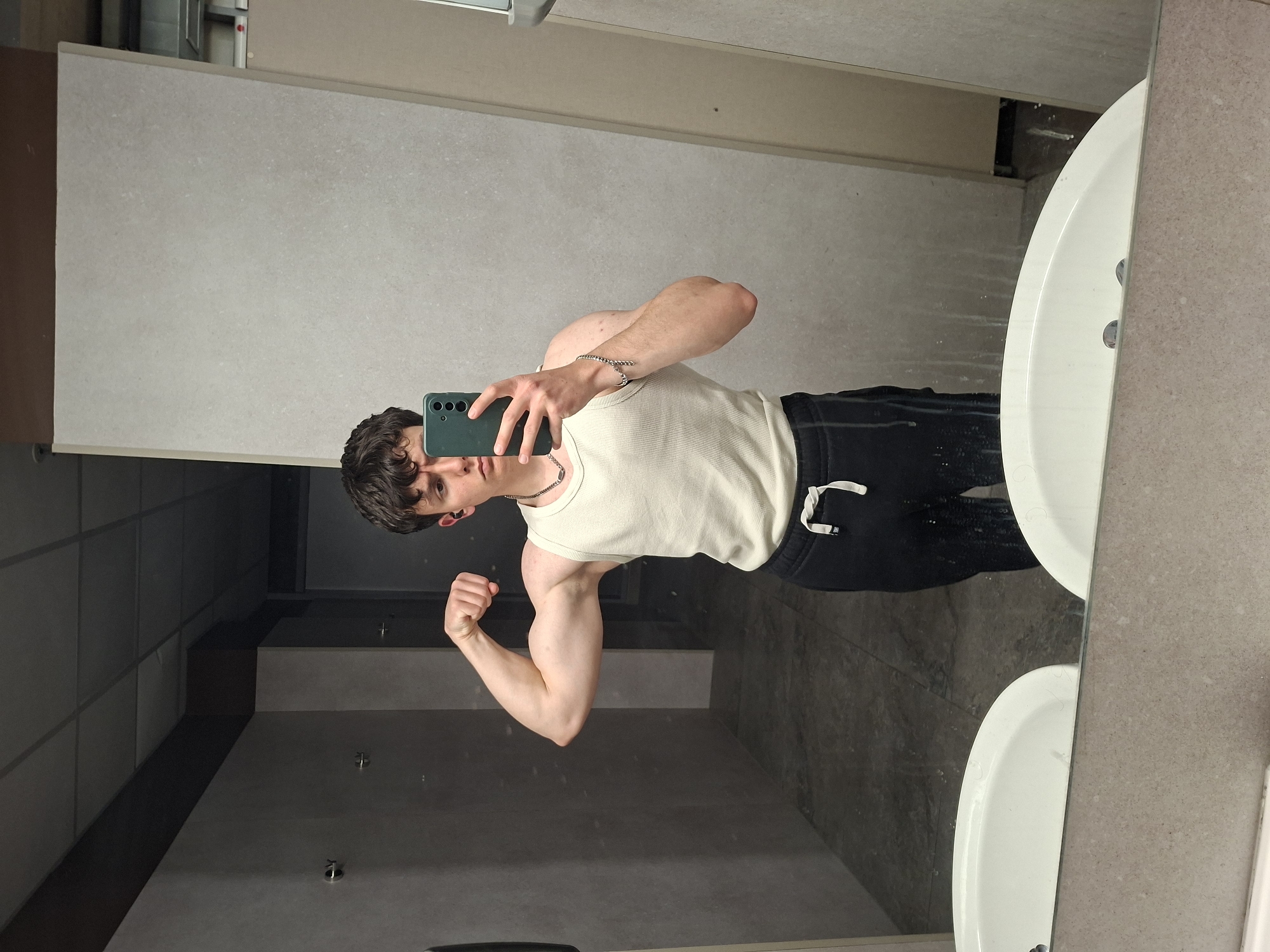A young adult male is visible in a mirror selfie, posing in what appears to be a public restroom. He is wearing a white tank top and black athletic pants, flexing his right bicep for the camera. The restroom has light gray wall tiles and a double sink vanity with white sinks. The background is nondescript, suggesting an institutional or commercial setting. There is no indication of the location being in Plovdiv, Bulgaria.