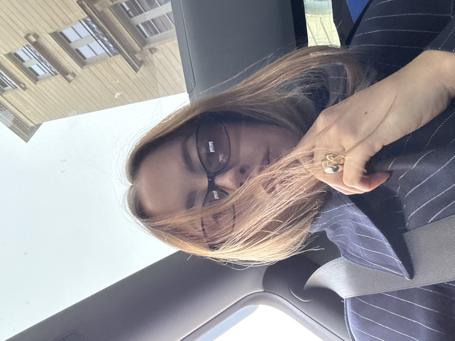 A forensic examination of the image reveals an individual inside a vehicle, located in the city of Unknown, Unknown. The subject is an adult female with medium-length, light brown hair, wearing dark-tinted oval sunglasses with black frames. Her face is partially obscured by hair and sunglasses. She is dressed in a dark pinstriped collared shirt or jacket over a darker garment. A light-colored seatbelt diagonally crosses her chest. Her left hand, adorned with a chunky gold-toned ring featuring an abstract design, is positioned near her lower face. The vehicle interior, visible on the left, is dark grey. Through the vehicle's window or sunroof, a light yellow/beige multi-story building with Haussmannian architectural elements, including windows and balconies with wrought iron railings, is discernible against an overcast sky. Specks are present on the glass surface.
