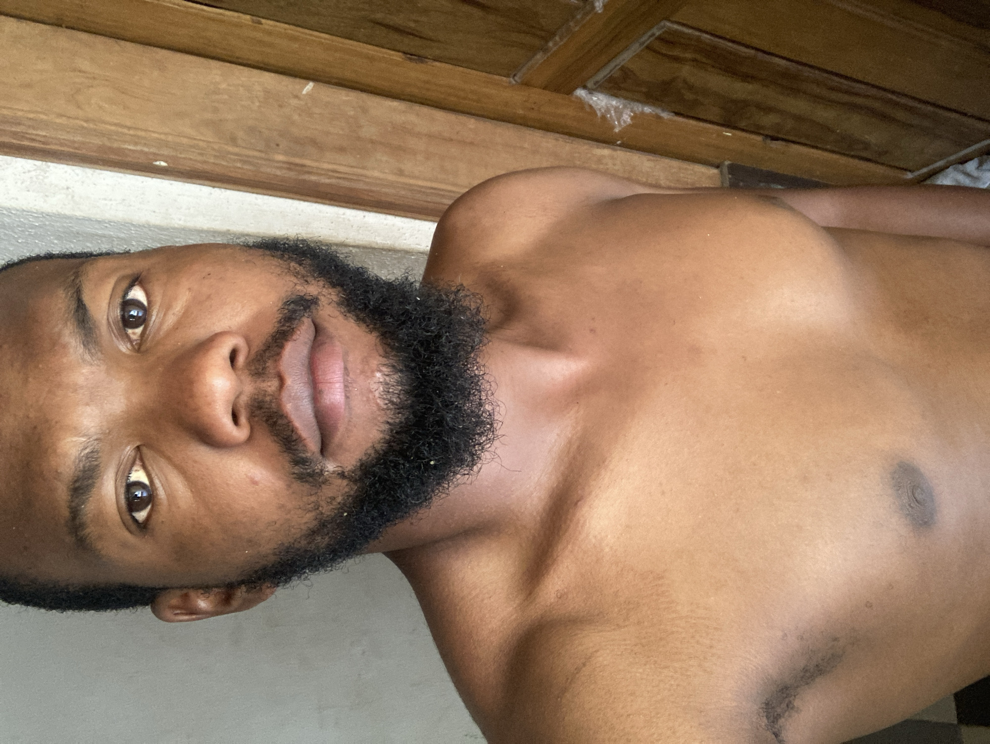 A man with dark skin and a full black beard is shown from the chest up, in a close-up, slightly angled shot. He is shirtless. The background appears to be an interior space with wooden panels above a light-colored wall. There is no clear information in the image to confirm that the location is in the city of Jos, Nigeria.