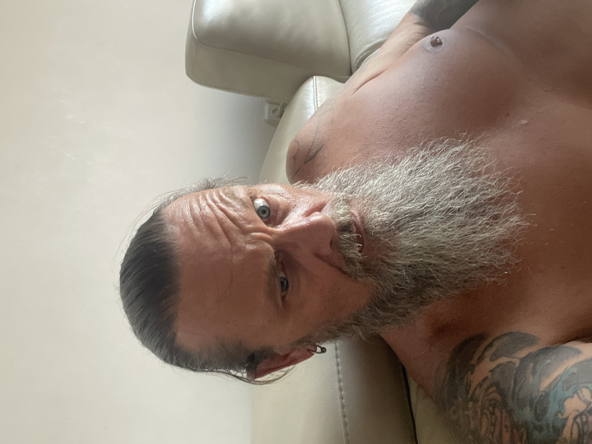 A close-up, chest-up selfie of a shirtless man with a long, grey beard and slicked-back dark hair is taken indoors, against a pale wall and a light-colored leather couch. The man has his hair pulled back into a ponytail and is wearing a silver hoop earring in his left ear. He has tattoos on his left arm and shoulder. His right arm is resting on the couch, and he is looking directly at the camera with a stern expression. The man's forehead is wrinkled, and his left eyebrow is raised. The lighting suggests it is daytime, possibly from a window to the right of the frame. There is no indication of the city of Netanya, Israel, within the image itself.