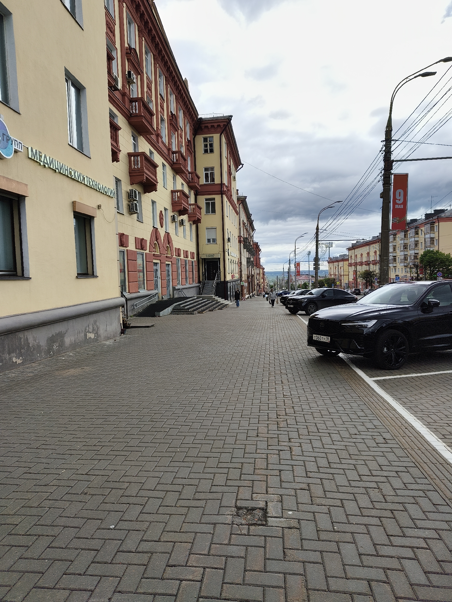 The image captures a street in Izhevsk, Russia. The view is along a brick-paved sidewalk on the left side of a street lined with cars. A black Volvo SUV is parked in the foreground, facing the viewer. Further down the street, there are several other parked cars. On the left side of the sidewalk, a building with a yellow facade is visible, displaying signage in Russian. Further down the street, there are more buildings of similar style. A banner with the date "9 MAY" hangs from a utility pole. The sky is overcast, suggesting cloudy weather conditions. A few people can be seen walking in the distance.