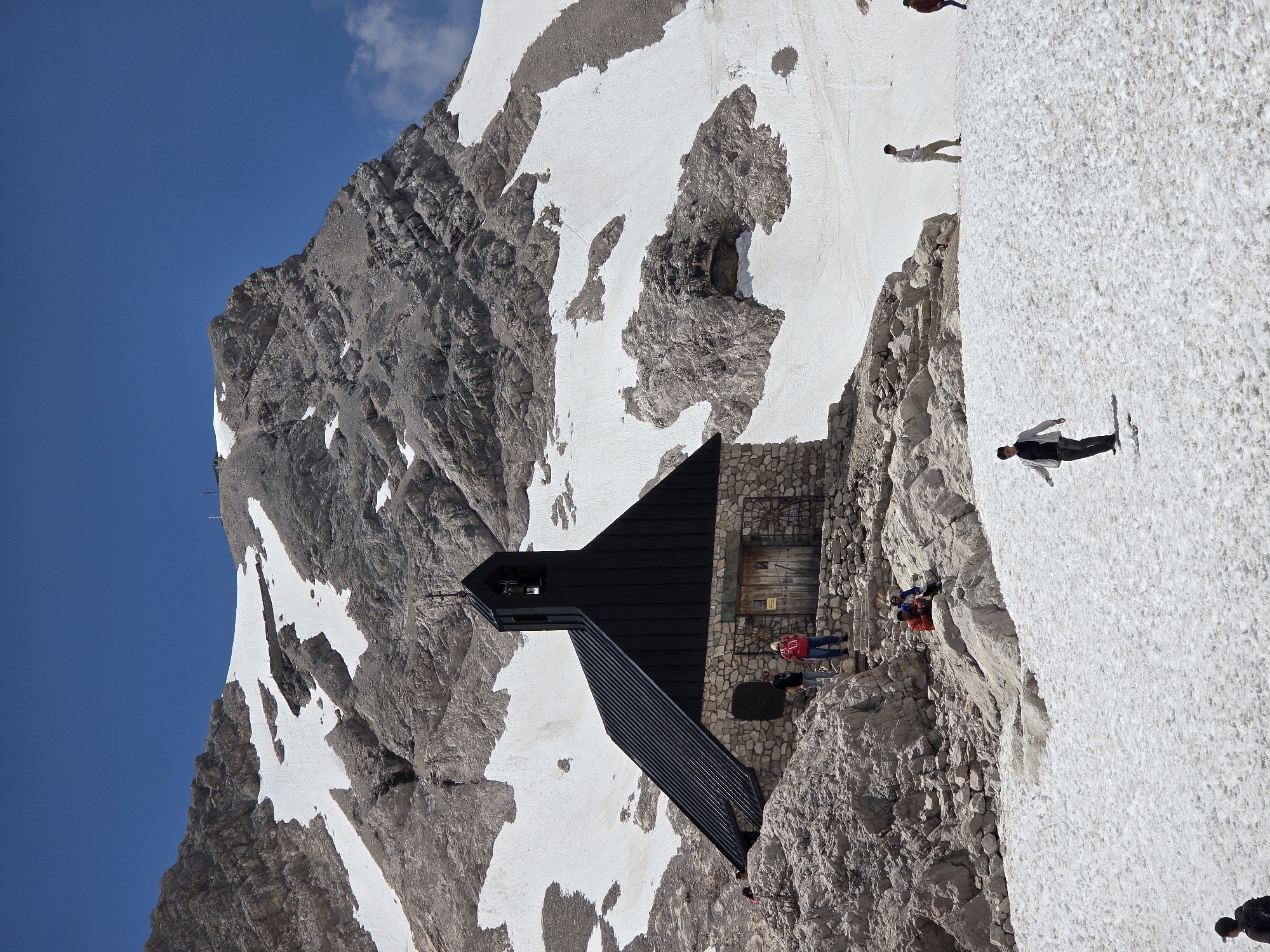The image depicts the summit area of the Zugspitze, Germany's highest mountain, located near Garmisch-Partenkirchen. A small stone building with a black, steeply pitched roof and a bell tower is the central subject, situated on a rocky outcrop. The surrounding landscape is mountainous, with significant patches of snow covering the slopes, indicative of a high-altitude environment.
Several people are visible in the scene. In the foreground, a person wearing a dark jacket and light-colored pants walks across the snow. Further up the slope, another individual in a light-colored shirt and pants is walking. Closer to the building, a group of people are gathered on the stone steps leading up to the entrance. Two individuals are standing by the building's wooden doors, one wearing a red shirt and the other a dark shirt. Two more people are seated on rocks to the left of the building. In the distance, the rocky, snow-dusted mountain peaks dominate the background under a clear blue sky. The overall impression is of a serene, majestic mountain landscape with human presence at a popular tourist destination.