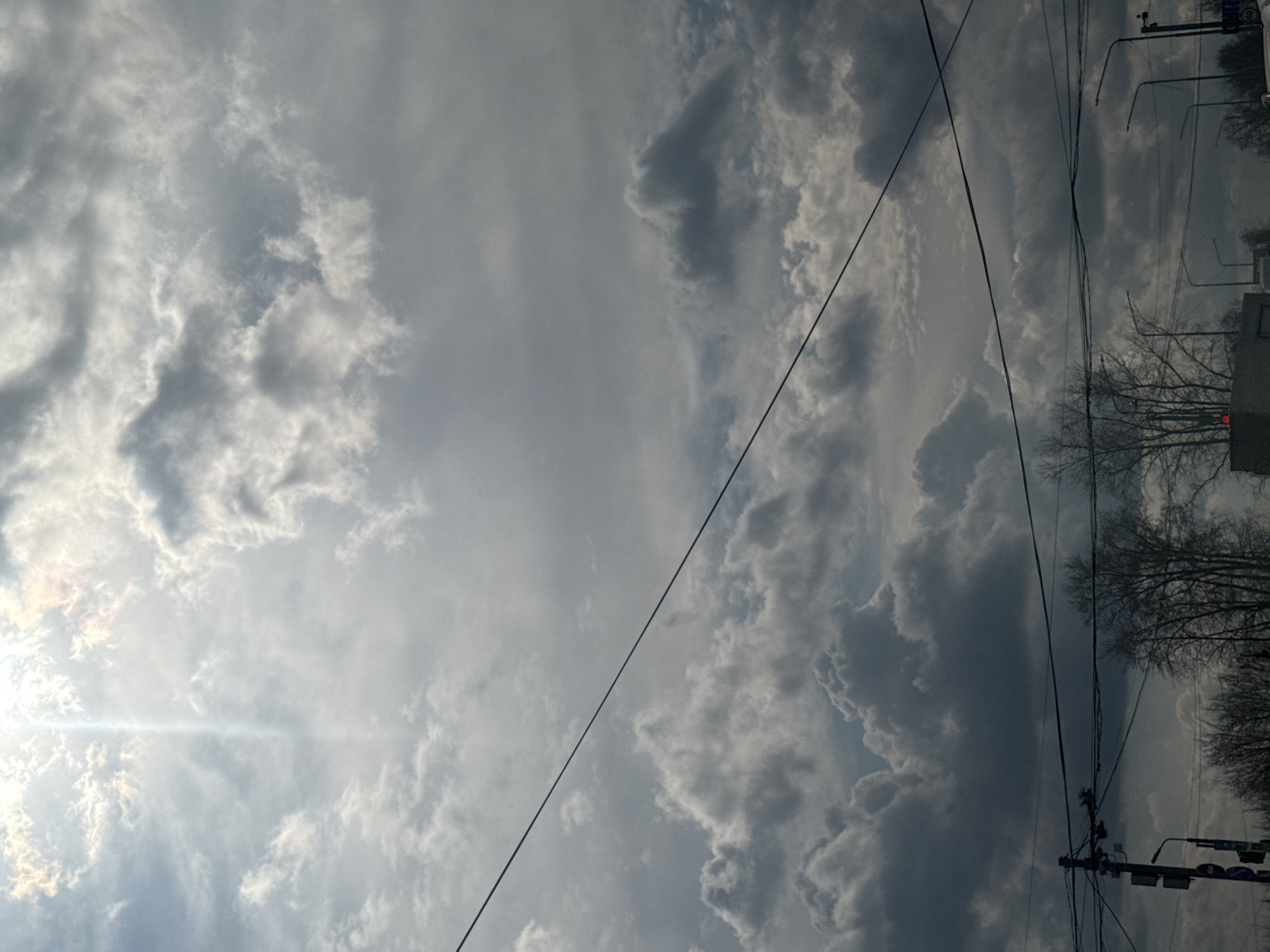 The image displays a sky filled with a diverse array of clouds, ranging from bright, sunlit white and light gray patches to darker, more ominous gray formations. On the upper left side, the sun is partially visible, creating a strong glare and a horizontal lens flare that stretches across the lighter clouds.
Several dark power lines or utility wires diagonally traverse the frame, particularly prominent on the right side, cutting across the cloudy backdrop. Towards the far right edge of the image, urban infrastructure is visible, including bare tree branches, suggesting a dormant, deciduous state typical of winter or early spring. Amidst the branches and wires, elements like utility poles, streetlights, and traffic signals can be discerned. A faint red light, possibly from a traffic signal, is visible near the bottom right corner.
The scene depicts an outdoor environment, likely an urban or suburban setting in Chaikovsky, Russia, during daytime. The weather appears partly cloudy with direct sunlight. No specific human activity is observable, and no discernible text is clearly visible on any signs or objects.