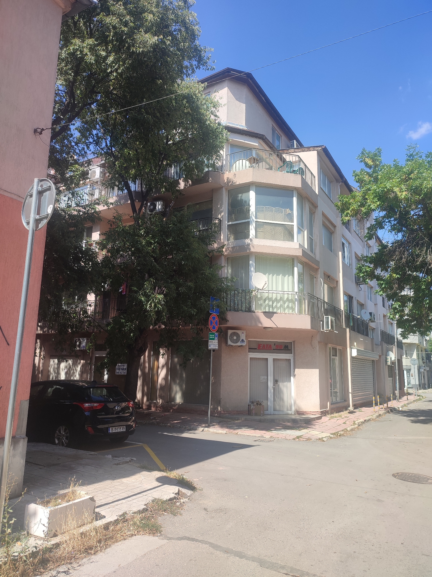 The image depicts a street view in Varna, Bulgaria, on a clear, sunny day. In the foreground, a black Hyundai hatchback is parked on the right side of a paved area with yellow markings. Adjacent to the parking area, a concrete planter box filled with dry grass and weeds is visible. A light pink building is partially visible on the left edge of the frame, with a silver street sign pole and a round traffic sign.
The main subject is a multi-story building with a light beige facade, featuring several balconies with black metal railings. The building has numerous windows, some with white curtains and satellite dishes attached. Air conditioning units are mounted on the exterior walls. A large tree with dense green foliage partially obscures the lower floors of the building. On the ground floor of this building, there is a shop with double glass doors and a roller shutter door. A sign above the glass doors reads "FAJA". Another sign attached to the wall of the building, above the parked car, has text in Bulgarian.
Further down the street, other buildings and shops are visible, as well as more trees and more parked cars. The street itself is paved with asphalt, and a manhole cover is visible in the road. The overall scene suggests a residential or commercial area in a city.