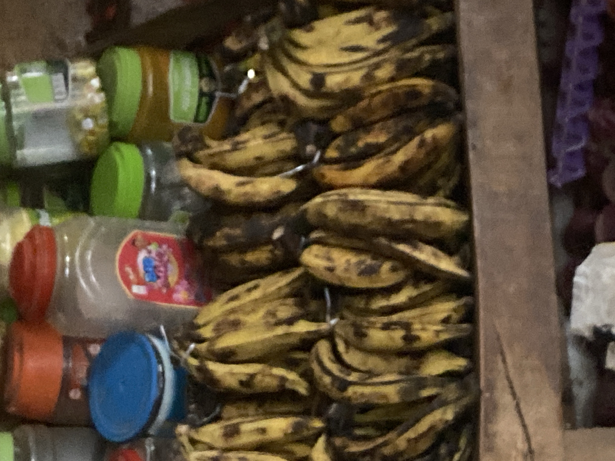 The image depicts a close-up, slightly out-of-focus view of a stall or market display featuring a bunch of ripe bananas in the foreground. The bananas are predominantly yellow with brown spots, indicating their ripeness. Behind the bananas, several containers are visible. To the left, there is a blue lid on a container. Further back, a large clear jar with a red lid, likely containing spices or grains, is prominent. To the right of this jar, there are several other clear jars with green lids, some appearing to contain yellowish or orange substances. The overall setting appears to be an outdoor or semi-enclosed market stall, common in cities like Lafia, Nigeria, where such produce and goods are displayed for sale. The lighting suggests it is daytime. No people are clearly visible or identifiable in the frame. The focus is on the produce and the containers in the background.