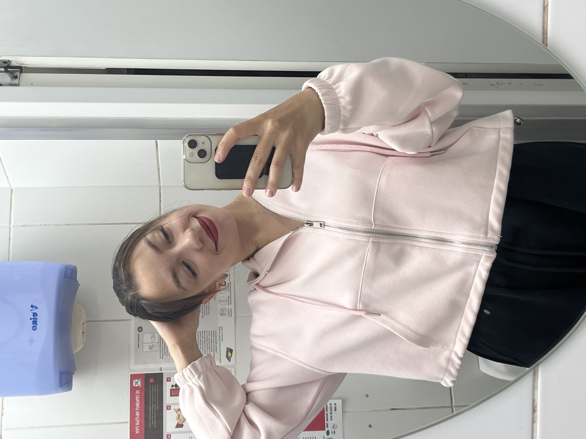 A young woman is taking a mirror selfie in what appears to be a public restroom, possibly in Chaikovsky, Russia. She is wearing a pale pink zippered hoodie and dark bottoms. The restroom has white tiled walls, a blue paper towel dispenser mounted on the wall, and a large circular mirror reflecting the woman and her surroundings. Instructional signs are visible on the wall behind her. She is holding a smartphone in her right hand and has her left hand on the back of her head, looking at her reflection with a slight smile. The lighting is bright, suggesting artificial light.