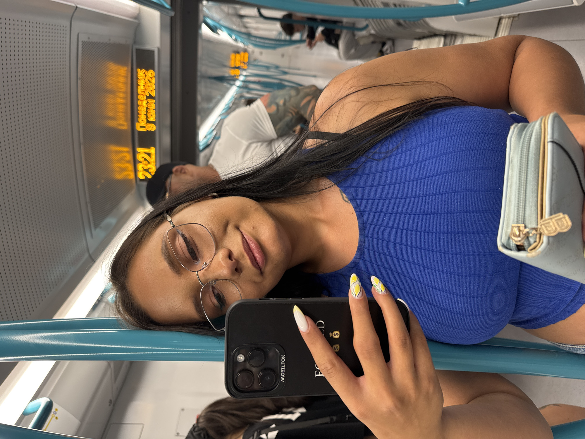 A young woman with dark hair and glasses takes a selfie inside a metro train. She is wearing a blue ribbed tank top and holding a black smartphone in her left hand. She is also holding a light blue coin purse with a gold zipper. In the background, other passengers are visible, as well as a digital display showing "23:21 Понеделник 8 Юни 2026" which translates to "23:21 Monday 8 June 2026". This indicates the image was taken in Sofia, Bulgaria.