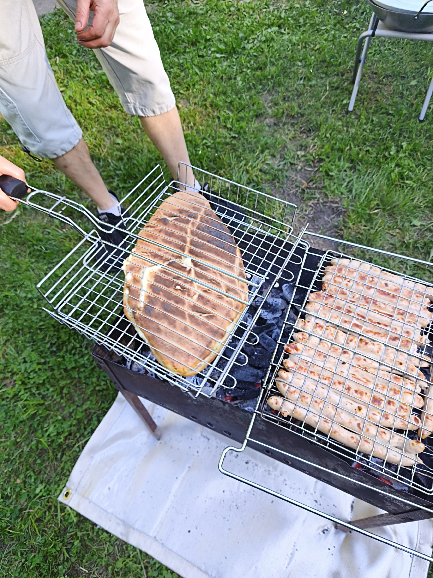 The image shows a person's legs and hands grilling food on a charcoal barbecue in what appears to be a backyard or park setting. The food being grilled consists of several rows of sausages on one side of the grill and a large, flatbread or loaf of bread on the other side, both in a metal grilling basket. The barbecue is placed on a dark cloth spread on green grass. In the background, a metal outdoor chair with a white pot is visible. The scene suggests an outdoor meal preparation activity.