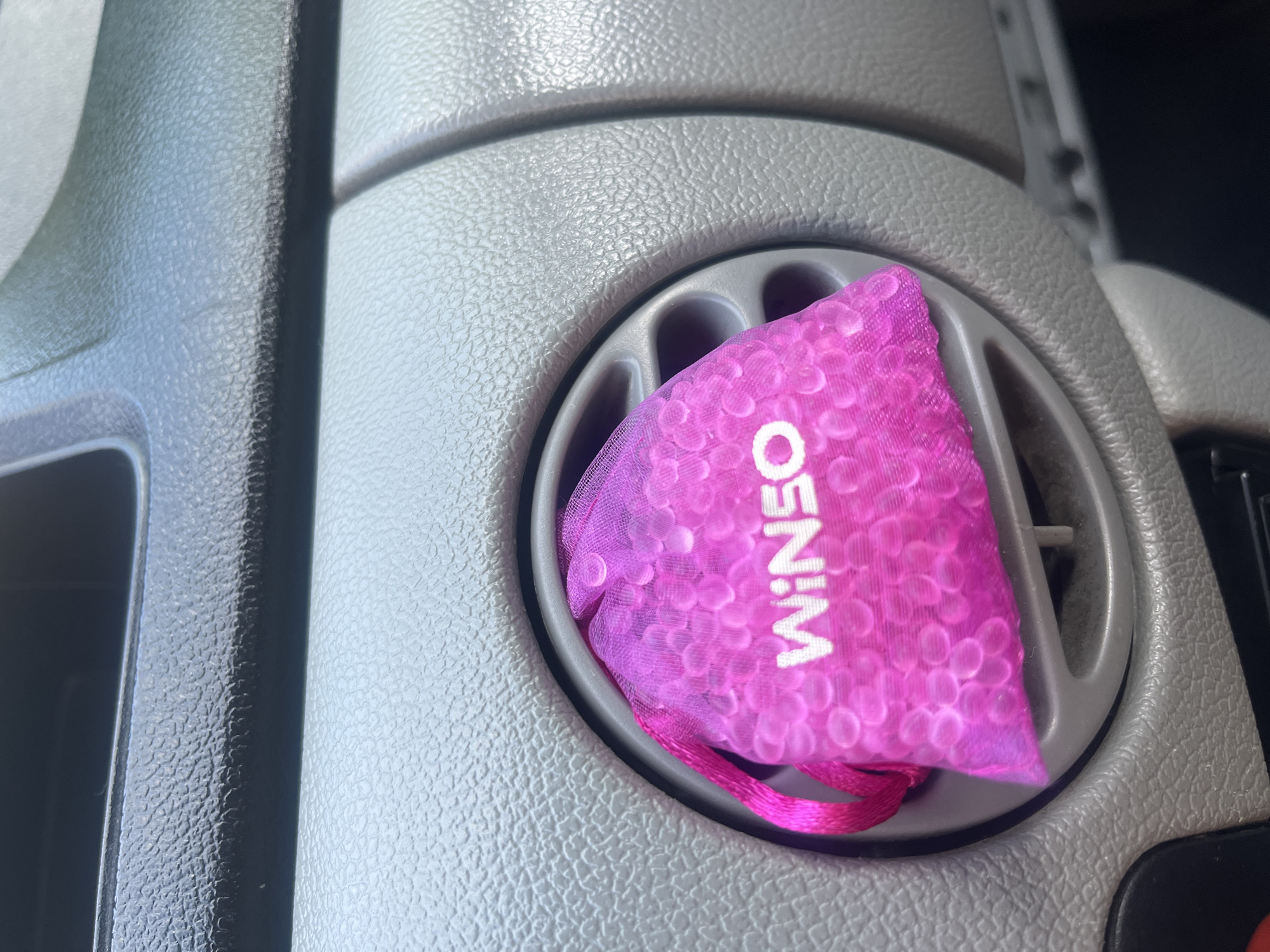 A pink mesh bag filled with small, round, pink beads is placed inside a car's air vent. The bag has a bright pink ribbon tie and the word "WINSO" is printed in white on the front of the bag. The interior of the car is visible, including the textured dashboard and the circular air vent. The overall lighting suggests the image was taken during the daytime, likely in a car parked outdoors. There is no indication of the city of Tarnivtsi, Ukraine, in the image.