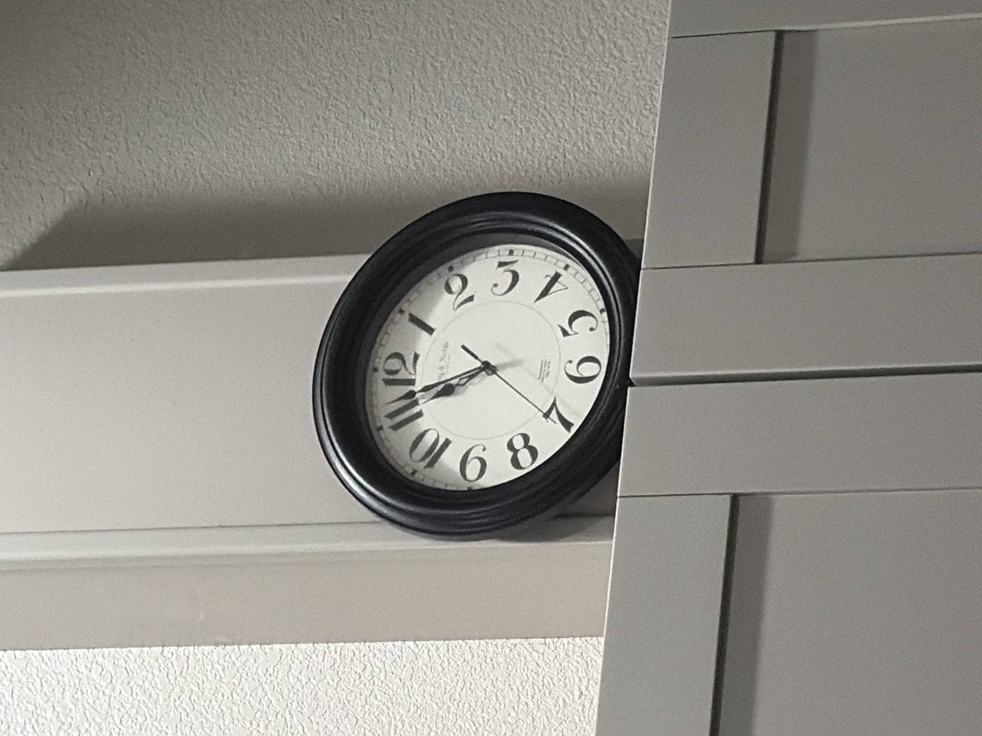 This is a close-up, eye-level shot of a black analog clock with a white face and black Roman numerals. The clock is positioned on a light-colored ledge, which is in front of a textured gray wall. To the right of the clock, a portion of a gray cabinet with horizontal paneling is visible. The clock hands are not clearly visible, making it impossible to determine the exact time. The lighting suggests it is daytime, though no specific time of day can be inferred. The environment appears to be indoors, possibly a home or office, given the presence of the clock and cabinet. There is no discernible text other than the numerals on the clock face. No people or weather conditions are visible. The image captures a simple, everyday object in a neutral setting.