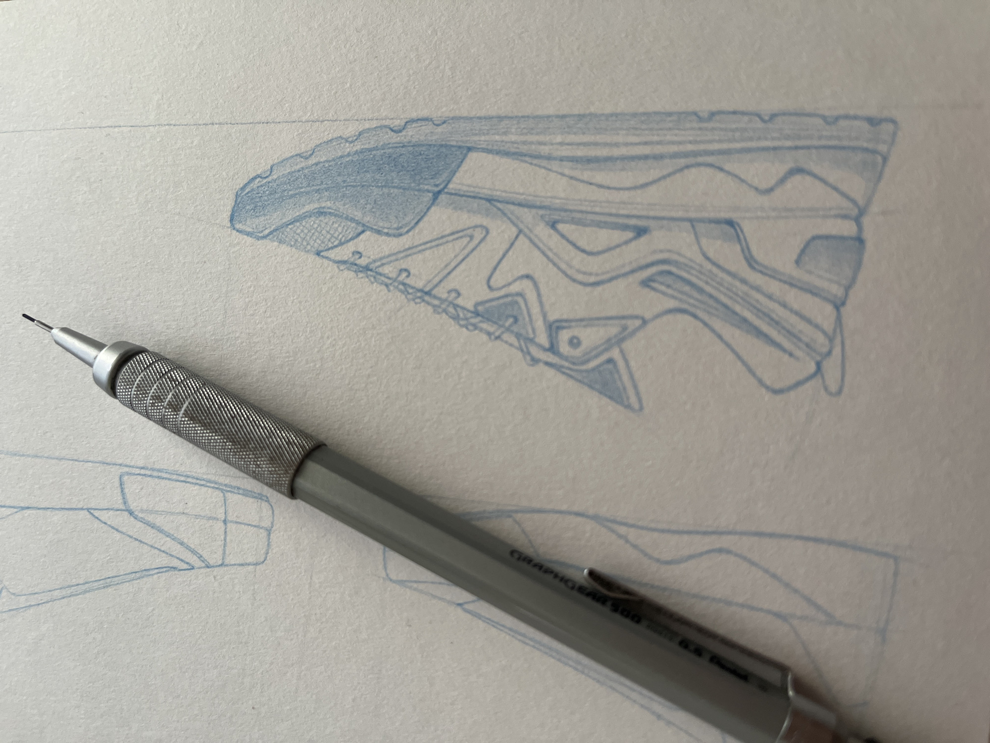 The image captures a close-up view of a detailed design sketch on white, textured paper, accompanied by a mechanical pencil. The primary subject is a blue pencil drawing of the side profile and sole of an athletic shoe or sneaker. This drawing is rendered with intricate lines and shading, depicting various layered components and patterns commonly found in footwear design.
A metallic gray mechanical pencil rests diagonally across the lower part of the paper. It features a distinct knurled grip near its extended lead, ready for use. Clearly visible text on the pencil identifies it as "GRAPHO GEAR 500 0.5 HB".
Faint blue lines from other preliminary sketches are visible on the paper in the foreground and background, suggesting an ongoing design process or multiple concepts. The scene is brightly lit, indicating an indoor environment, likely a desk or studio. The image conveys a sense of precision, creativity, and focus on industrial design. While the location context of Krasnoyarsk, Russia, is provided, the visual elements within the image do not offer any specific geographical indicators.