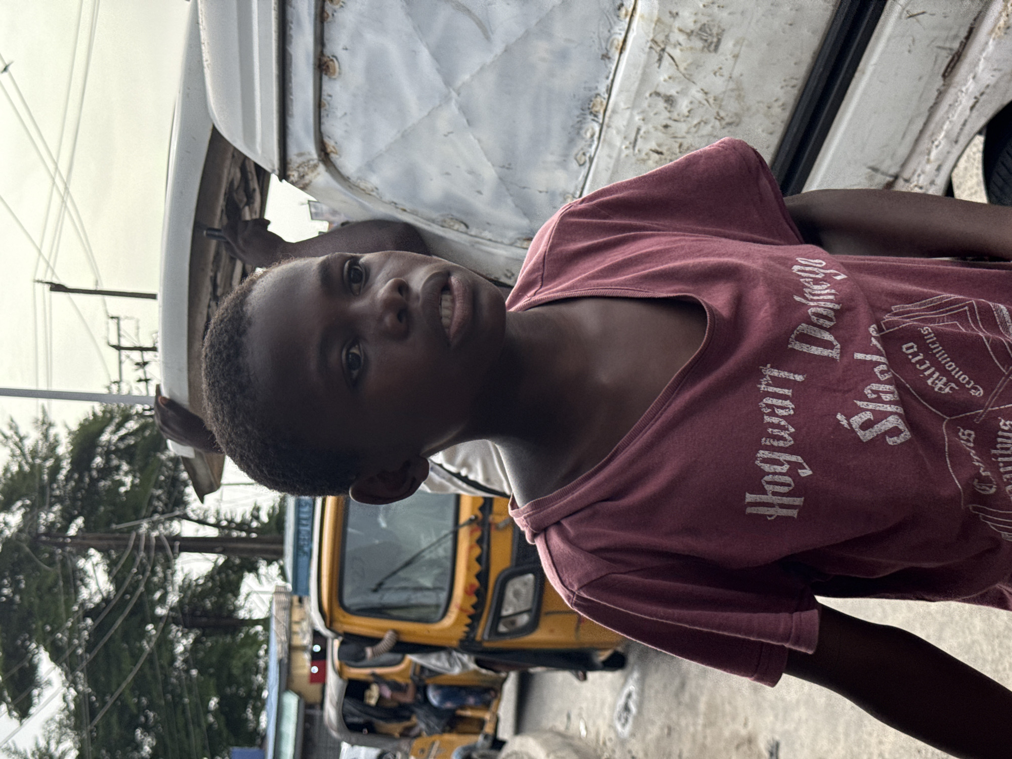 A young boy in a maroon "Hogwarts School" t-shirt stands in the city of Ado, Nigeria. He is positioned near a white, weathered vehicle. Behind him, a yellow tricycle taxi is visible, along with other people in the background. Overhead power lines are present. The setting appears to be an urban street.