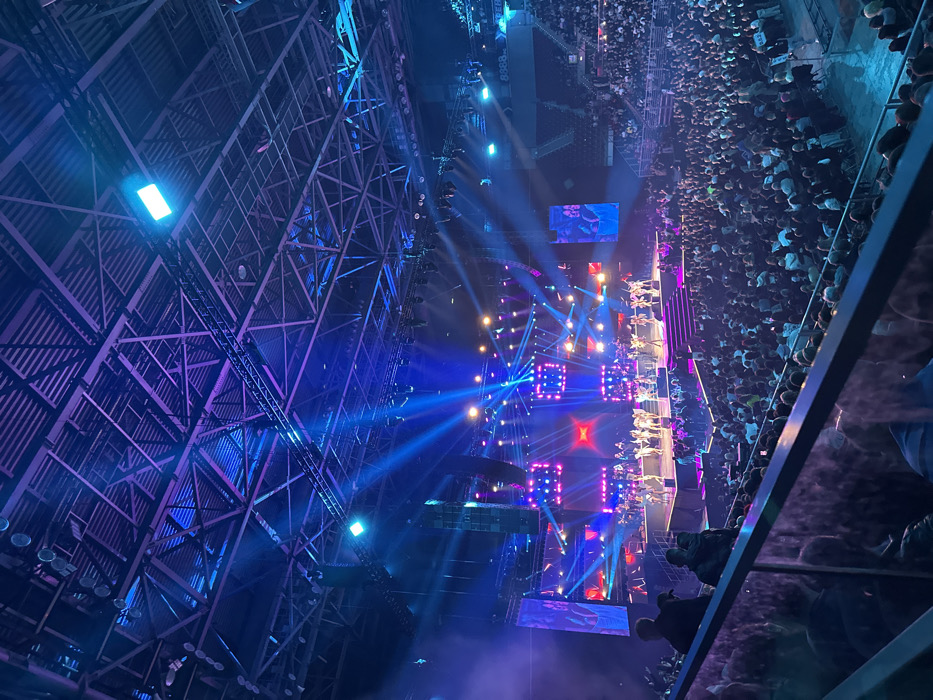 The image captures an expansive view of a live performance within a large indoor arena, identified as Arena Armeec (now Arena Sofia) in Sofia, Bulgaria. The perspective is from an elevated spectator section, looking down towards the stage and a densely packed audience.
The arena's ceiling features a robust, exposed steel truss structure, painted dark, from which numerous professional lighting fixtures, rigging, and large line array speaker systems are suspended. These structural elements are illuminated by ambient blue and purple stage lighting.
The main stage is positioned at the far end of the arena floor, characterized by multiple large LED screens or light panels displaying dynamic graphics in red, blue, and purple hues, centered around a prominent 'X' motif. Approximately 20 to 30 performers, clad in light-colored costumes, are actively engaged in a synchronized routine across various platforms on the stage. Intense, multi-directional beams of light in shades of blue, purple, magenta, and white emanate from fixtures, cutting through atmospheric haze above the stage.
The arena floor and lower tiered seating areas are filled with a large crowd of several thousand spectators, all oriented towards the stage. In the immediate foreground, a metal and transparent barrier, characteristic of an upper seating level, is visible, with the silhouettes of a few individuals observing the performance. A blue sign displaying the number "8388" is partially visible on the right side of the arena, above the audience seating.
