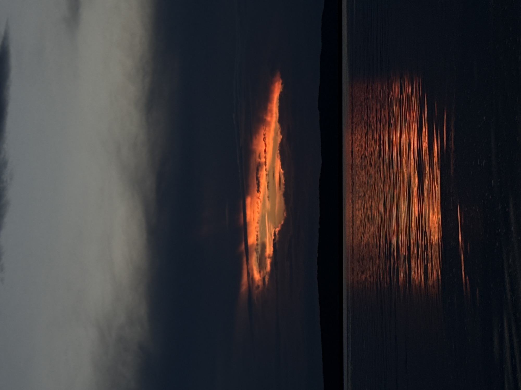 This is a photograph taken at sunset in the city of Chaikovsky, Russia. The image depicts a body of water, likely a river or reservoir, with a dark, silhouetted treeline visible on the horizon. Above the treeline, the sky is dominated by dark, brooding clouds. However, a striking break in the clouds reveals a vibrant band of orange and red light, suggesting the last rays of the setting sun are piercing through. This dramatic light is reflected in the water, creating a shimmering trail of color across its dark surface. The overall mood is serene yet dramatic, capturing the fleeting beauty of twilight. There are no people or man-made structures clearly visible in the frame.