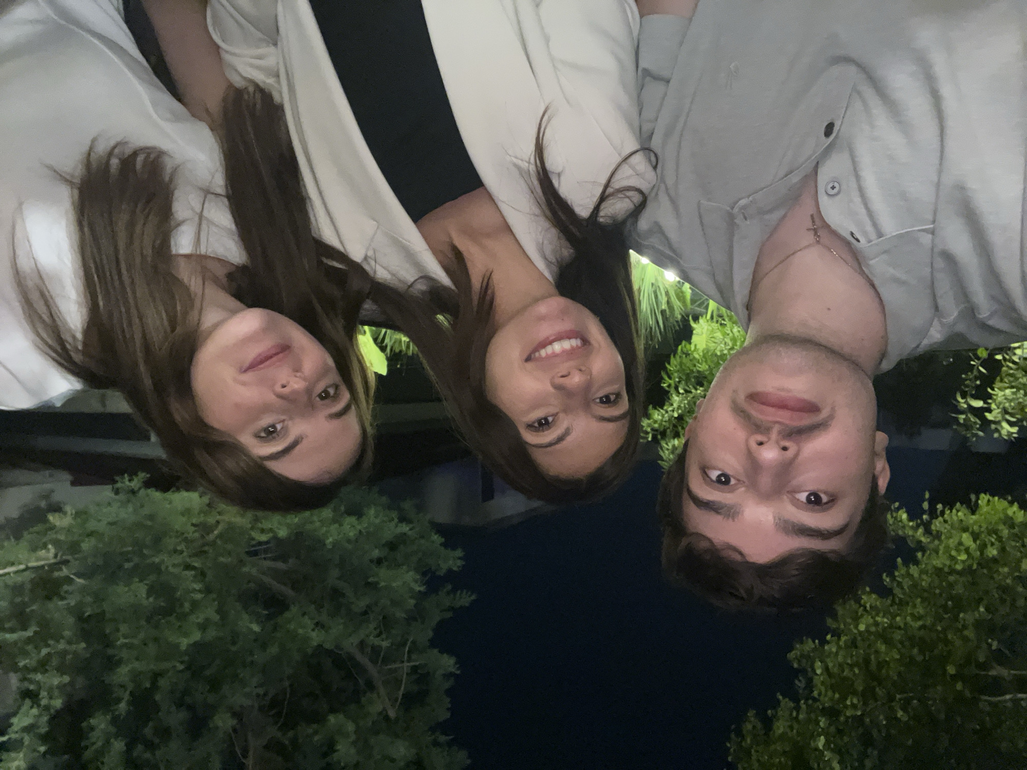 Three individuals are posing for a photograph at night, seemingly in San Bernardino Tlaxcalancingo, Mexico.
The individual on the left is a female with long brown hair, wearing a white top. The person in the middle is also a female with long brown hair, wearing a white blazer over a black top. The individual on the right is a male wearing a light grey button-down shirt with a cross necklace visible.
They appear to be outside with trees visible behind them. The lighting suggests it is nighttime.