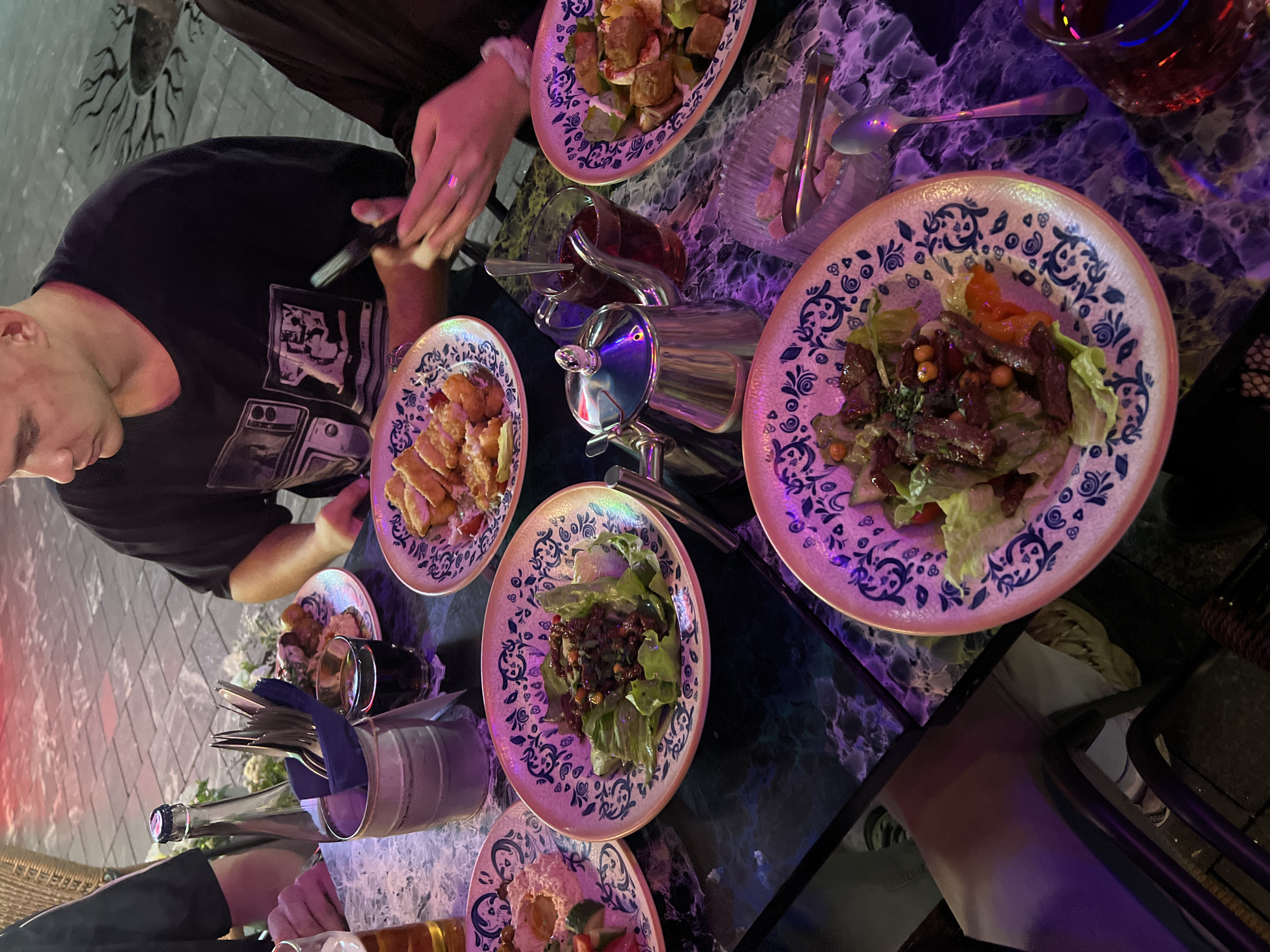 This image captures a dining scene, likely outdoors, on a table with a dark, marbled surface and a blue and white patterned edge. The lighting is ambient, with hints of purple and red, suggesting nighttime or evening. The ground visible in the background appears to be paved with rectangular stones.
In the foreground, several plates of food are arranged. One plate holds a salad with lettuce and what appears to be sliced meat, garnished with small orange beads. Another plate contains a salad with cubed ingredients and a reddish dressing. A third plate features fried, breaded food items, possibly chicken. Further back, a plate shows a dip, possibly hummus, with cucumber and tomato slices. A silver teapot with a red liquid inside, likely tea, sits on the table alongside a small glass also containing the red liquid. Cutlery is present, including forks and spoons, some in a metal bucket with a dark blue napkin. A glass of amber liquid, likely beer, is also visible.
Three individuals are partially visible. On the left, a person's arm and hand are seen holding a glass. In the center, a man wearing a black t-shirt with a graphic print is seated, looking downwards. His right hand is resting on the table. To the right, another person's hands are visible, one holding a dark-colored smartphone and the other gesturing near the phone, possibly interacting with it. A ring is visible on one of the fingers.
The presence of a smartphone, the style of the tableware, and the paved outdoor setting suggest a casual dining establishment. Without further context or distinct landmarks, pinpointing the exact location within Nizhny Novgorod, Russia, is not possible based solely on the visual information provided. However, the overall ambiance suggests a restaurant or cafe setting.