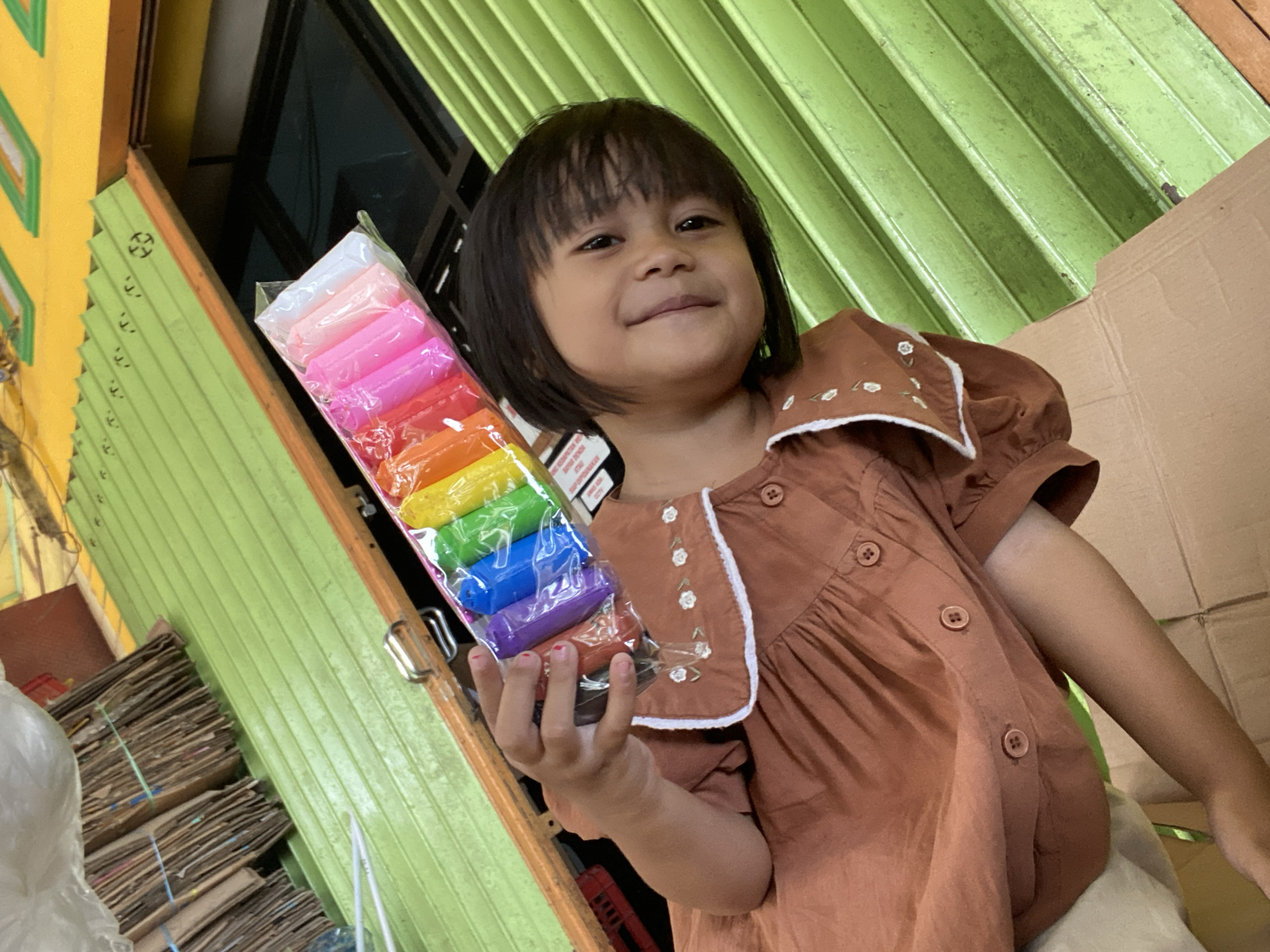 A young girl with dark hair smiles at the camera, holding a package of colorful playdough sticks in her left hand. She is wearing a brown dress with white trim and small floral embroidery. The girl is positioned in front of a green, corrugated metal shutter. To her left, stacks of cardboard are visible. To her right, a cardboard box is partially in frame. The scene appears to be outdoors, possibly in front of a shop. The lighting suggests it is daytime. The girl's expression conveys happiness. There is no visible text.