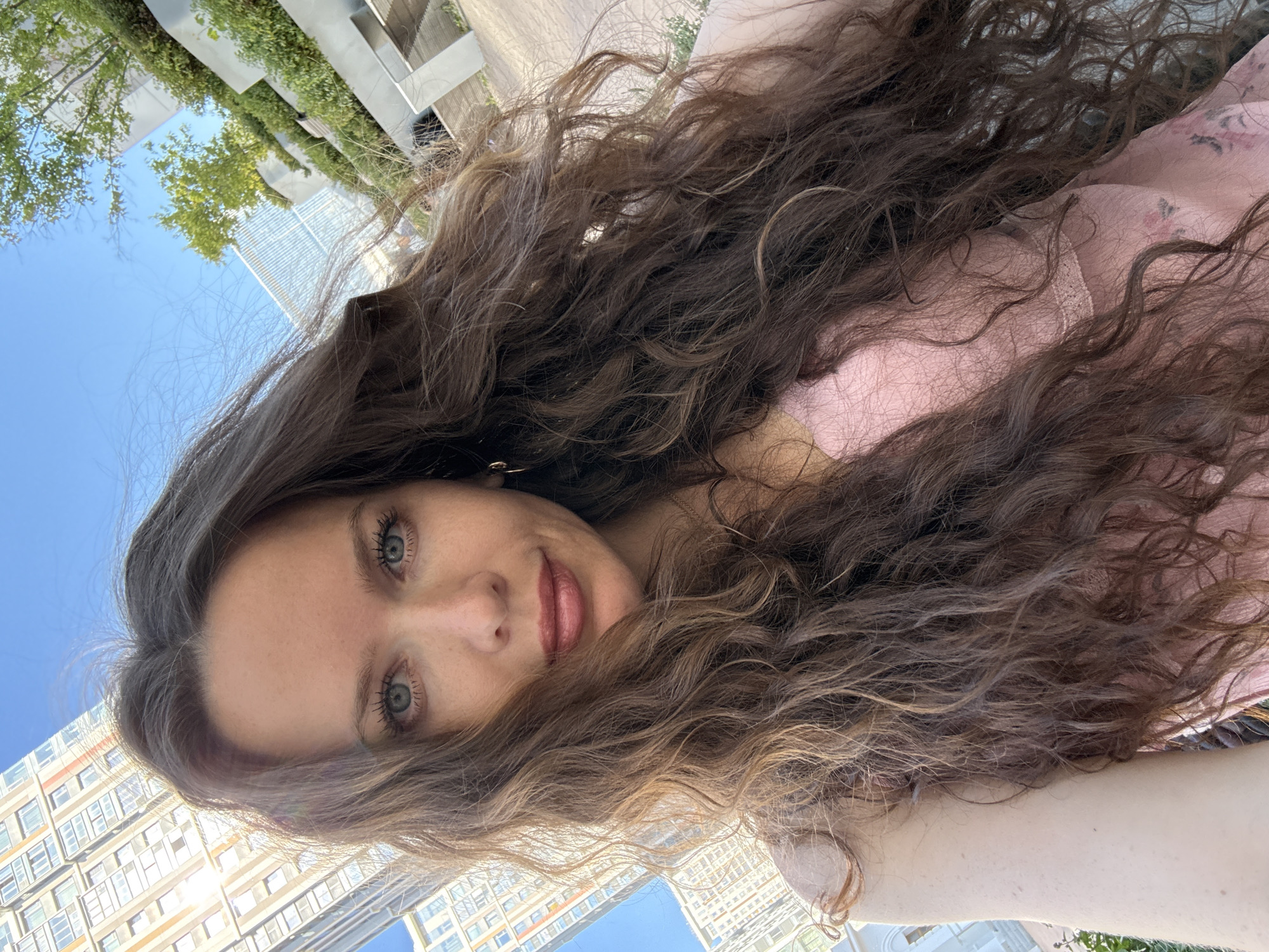 A woman with long, wavy brown hair, blue eyes, and fair skin takes a selfie outdoors. She is wearing a pink top with a floral pattern and a lace trim. Her hair is blowing in the wind. In the background, there are tall buildings, trees, and a blue sky. The architecture suggests an urban environment, possibly a modern residential area or business district. The presence of lush greenery, possibly in a landscaped urban park or terrace, further points to a city setting. The specific architectural style of the buildings, characterized by their height and modern design, along with the urban park elements, is consistent with many newer developments found in Moscow, Russia.