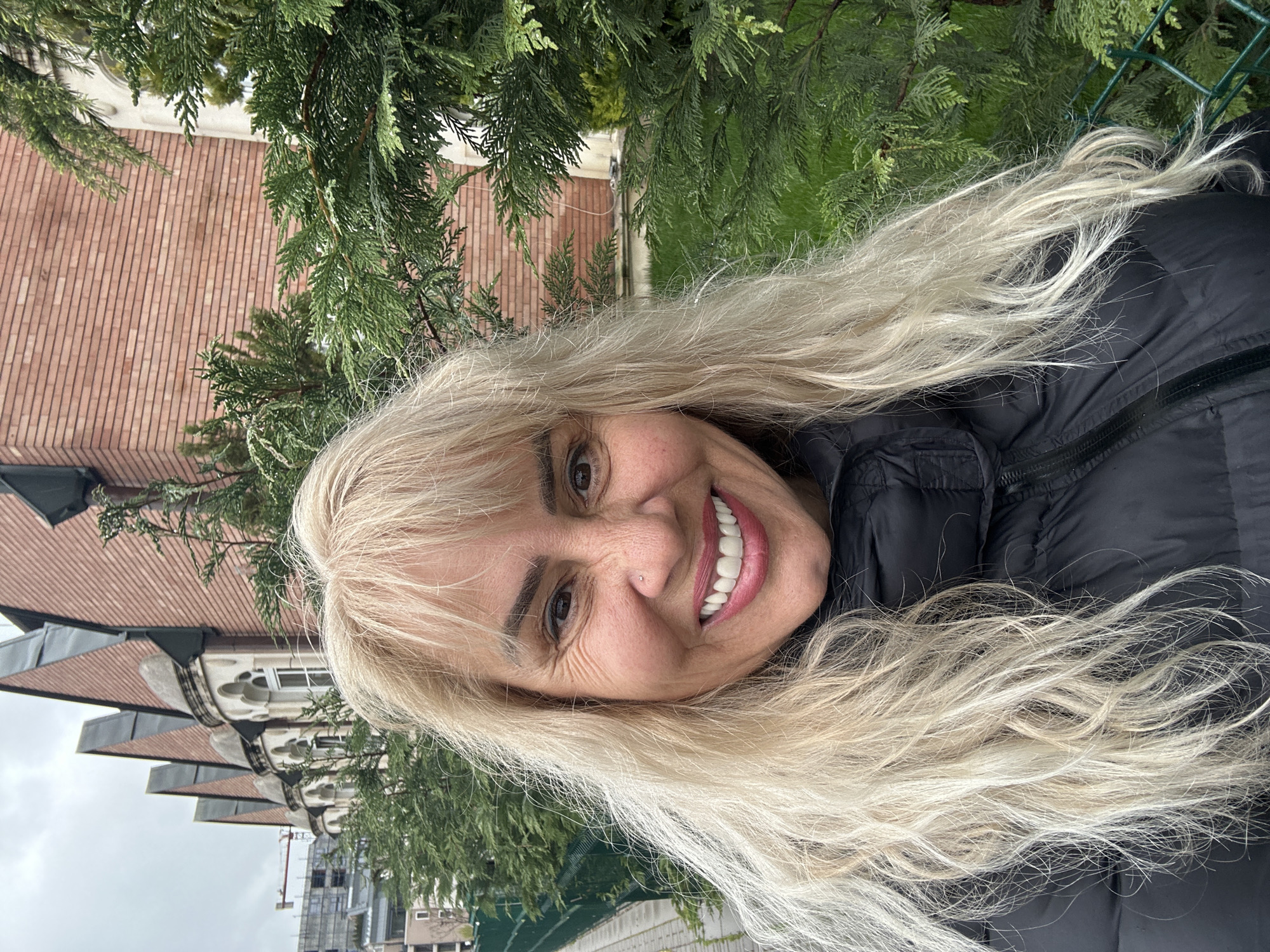 A woman with long blonde hair is smiling at the camera. She is wearing a black jacket. Behind her are green trees, a red brick building, and in the distance, construction cranes and buildings. The image appears to have been taken outdoors, in Sofia, Bulgaria.