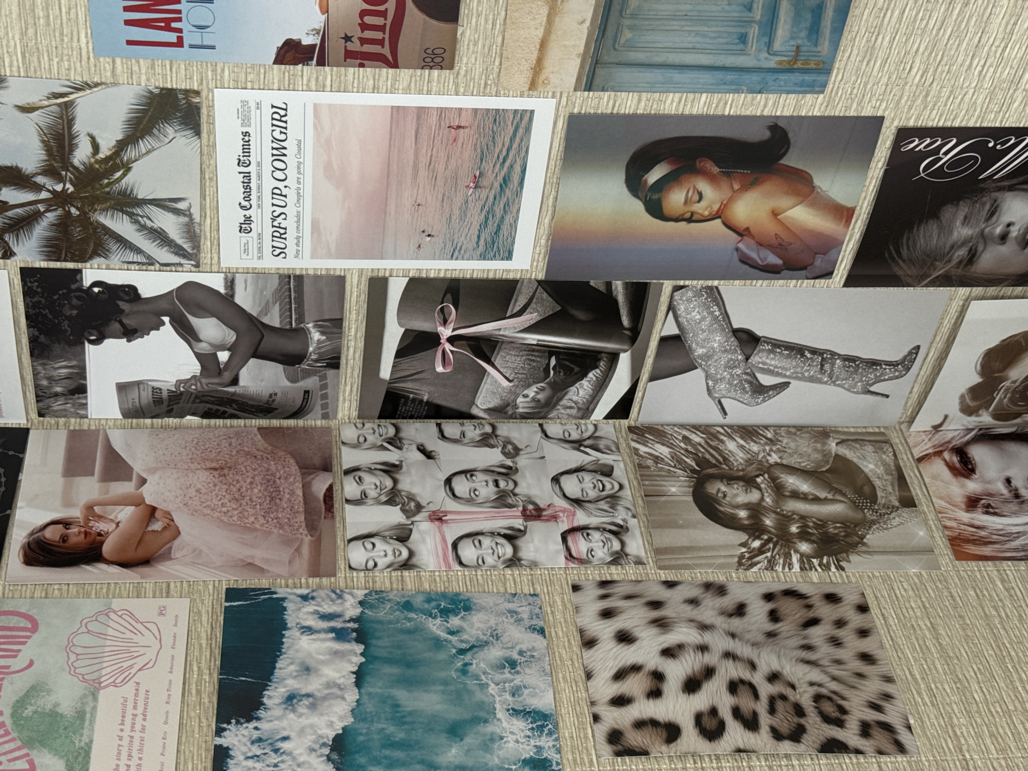 The image shows a collection of diverse postcards and images arranged on a surface with a wood-like texture. The postcards and images include a palm tree, a newspaper titled "The Coastal Times" with the headline "Surf's Up, Cowgirl", an illustration of a woman reading, a portrait of Ariana Grande, a magazine cover with "Rae" written on it, a blue door, a postcard for a story about a mermaid, ocean waves, a cheetah print, images of women in various settings, and shiny boots. The items are presented close-up, occupying the entire frame. There is no evidence to suggest this collection is in the city of Sochi, Russia.