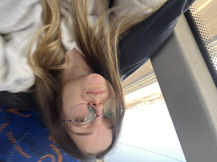 A young Caucasian female is depicted in a selfie, taken from the chest up, inside a vehicle, located within the city of Zaozerye, Russia. She has long, wavy, light brown hair with blonde highlights and wears circular, thin-rimmed glasses. Her facial expression is neutral as she looks directly towards the camera. She is wearing a light-colored, textured top, with a dark garment visible on her left side. To her left, a gray, textured window frame with a perforated section is visible, revealing an exterior view. Outside the window, multiple parallel train tracks are visible under a bright, clear sky, with light brown ground and indistinct distant structures or vegetation. On the far right of the frame, a blue seat with a darker abstract pattern is partially visible. The scene is illuminated by natural daylight, indicating daytime.