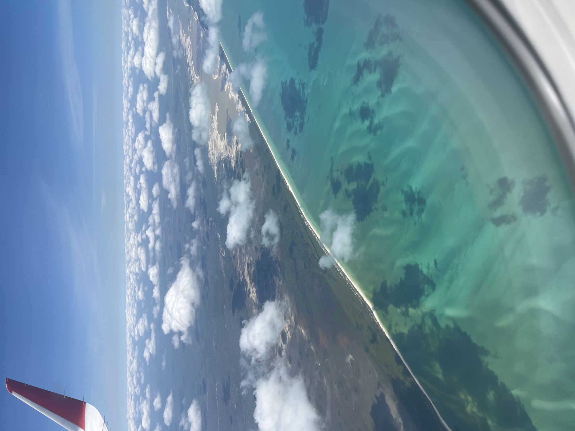 This media file presents an aerial view of a coastal landscape, captured from the window of an airplane during daytime. The scene is bisected by a distinct coastline. On the left, a bright blue sky is adorned with numerous scattered white cumulus clouds, some casting soft shadows.
Below the clouds, the landmass stretches along the coast, featuring a narrow, pristine white sand beach that separates it from the ocean. The land itself is a mosaic of dark green vegetation, likely coastal forests or mangroves, interspersed with lighter, possibly sandy or scrubland areas.
To the right of the coastline, a vast expanse of turquoise water dominates, indicating clear, shallow conditions. Intricate patterns are visible beneath the water's surface, suggestive of sandbars, reefs, or seagrass beds. Darker patches within the water are present, likely shadows from the overhead clouds or deeper marine topography.
In the lower left corner, a portion of a red and white airplane wingtip is visible, partially framing the view. The curved edge of an airplane window frame is also visible along the right side of the image. The weather appears clear and sunny. There is no visible text in the image. The location, consistent with Xuilub, Mexico, showcases a beautiful tropical or subtropical coastal environment.