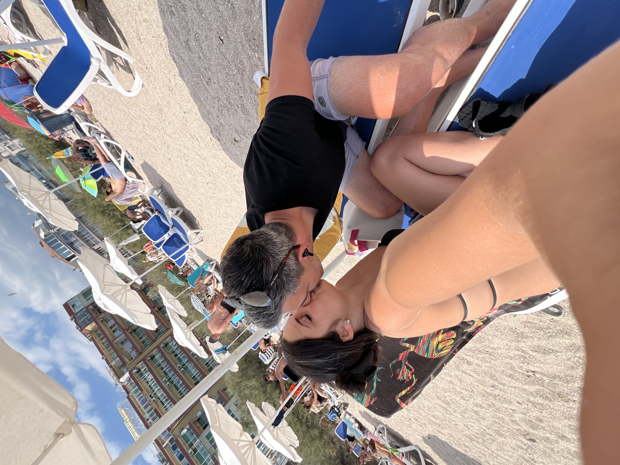 This is a candid, eye-level, close-up shot taken on a beach in Pomorie, Bulgaria, featuring a couple kissing. The woman, in the foreground and to the left, has dark hair pulled back and is wearing a black bikini top. Her eyes are closed, and she has a stud earring in her left ear. The man, to her right, has graying hair and is wearing sunglasses on top of his head and a black t-shirt. He is leaning in to kiss the woman.
They are both lying on blue lounge chairs on a beach with light-colored gravel. A colorful towel with an abstract design is draped over the woman's chair. In the background, there are other beachgoers, white umbrellas, and a multi-story hotel with a red facade. The sky is blue with some clouds, and a bird is visible in the upper part of the image. The overall scene depicts a romantic moment on a sunny day at the beach.