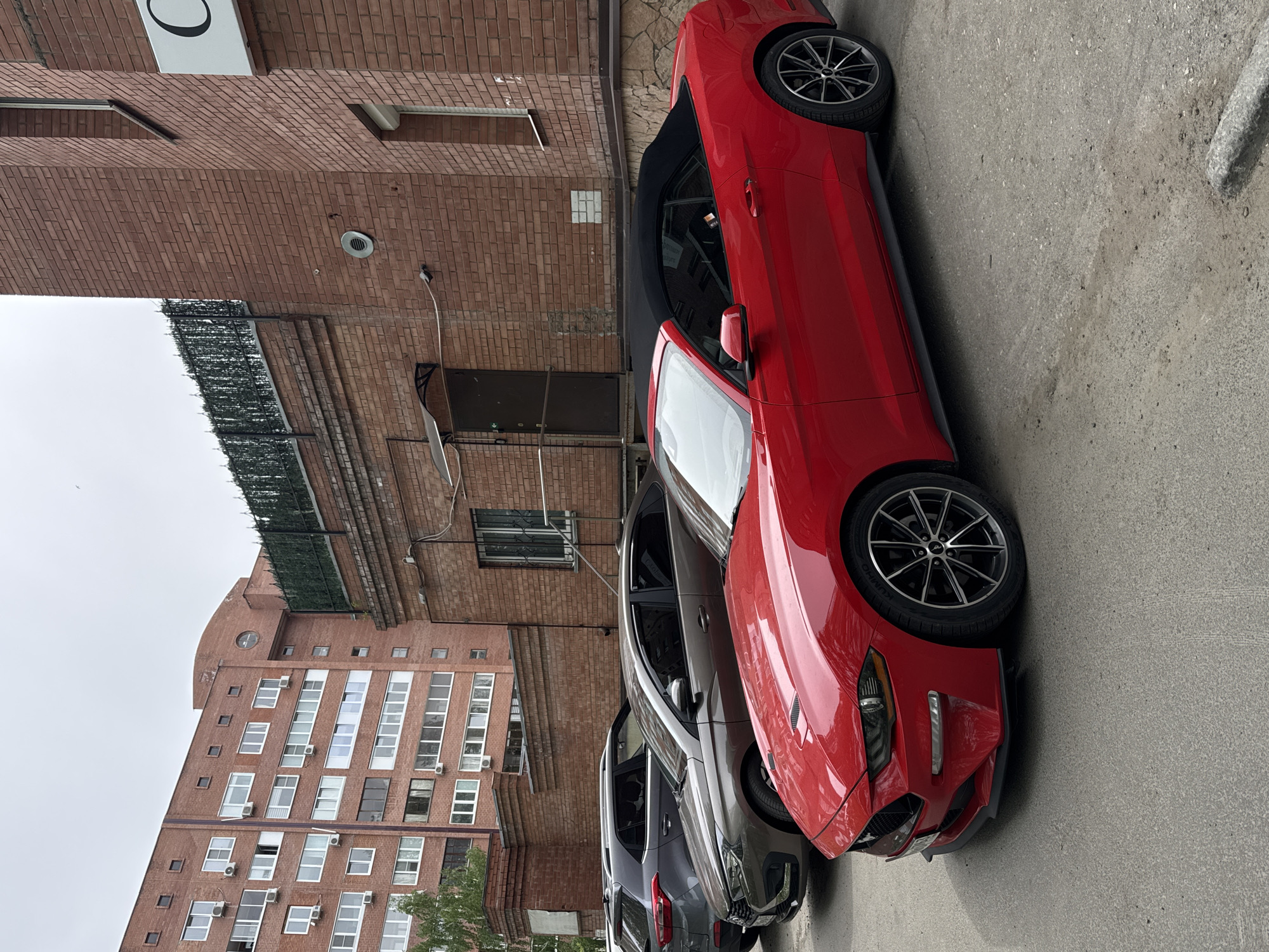 The image depicts a red convertible car parked on an asphalt street in an urban environment. The vehicle is positioned adjacent to a building constructed of red brick. Nearby are additional parked vehicles. In the background, a taller building of similar brick construction is visible. The sky is overcast, and the overall impression is one of a cloudy day in the city.