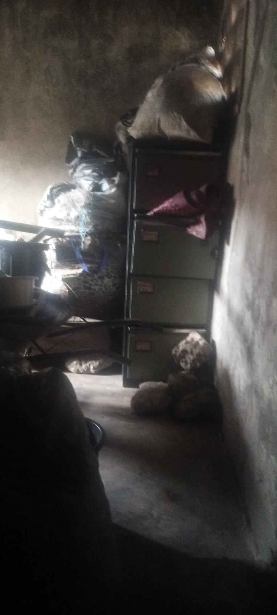 The image shows an interior scene in Sabon Tasha, Nigeria. A metal filing cabinet with three drawers stands against a concrete wall. Bags and other items are stacked on top of the cabinet. An object covered in a leopard print is to the left of the cabinet. There are several rocks or stones on the floor in front of the cabinet. The foreground is dark.