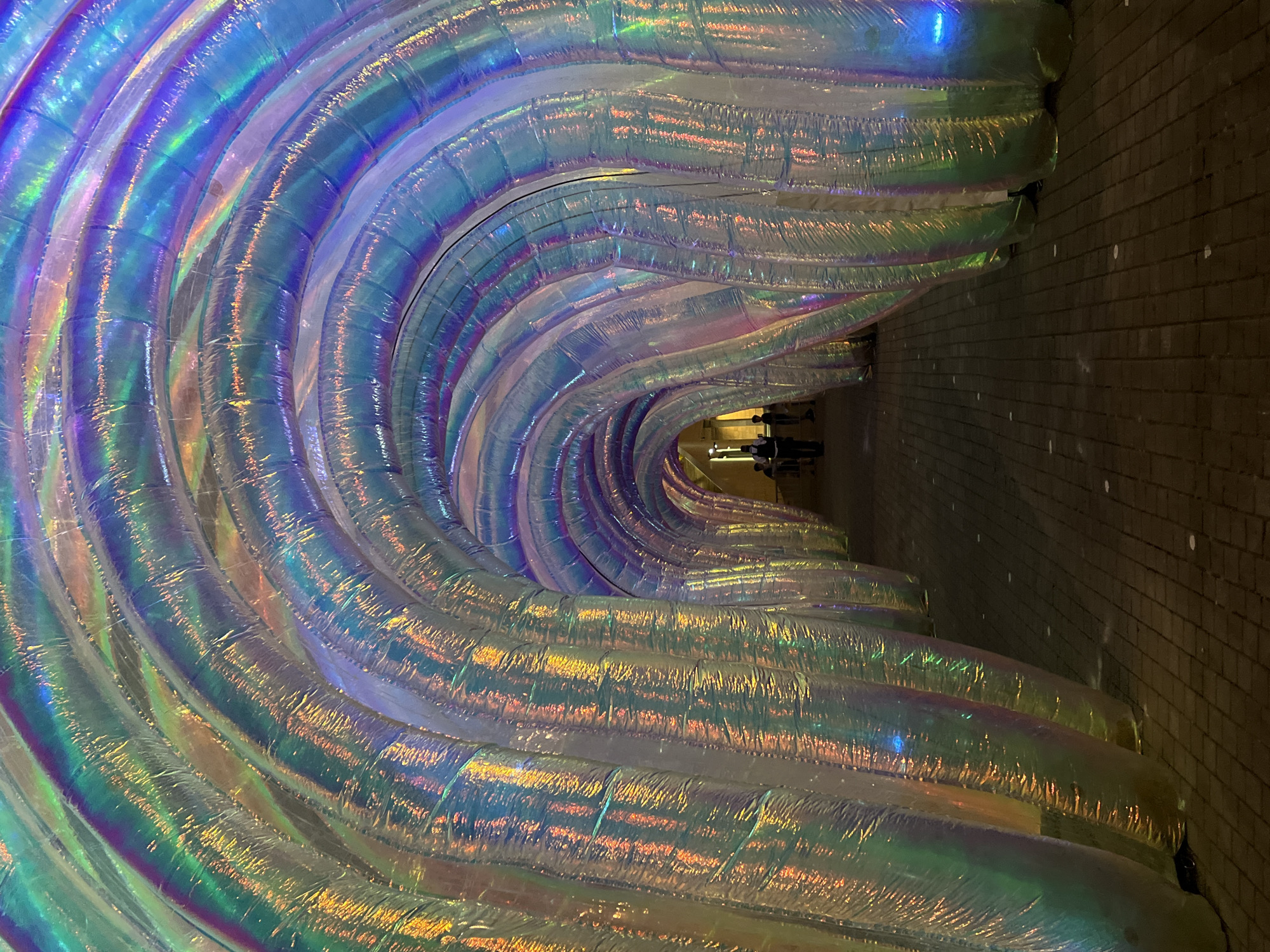 The image shows a tunnel-like structure with iridescent, inflated tubes arching overhead. The material of the tubes reflects light in a rainbow of colors, including blue, green, purple, and yellow. The ground is paved with dark bricks. In the distance, at the end of the tunnel, several people are visible, silhouetted against the brighter light at the far end. The overall impression is one of an art installation, likely in an outdoor public space. This installation is located at the Marina of Bilbao, Spain.