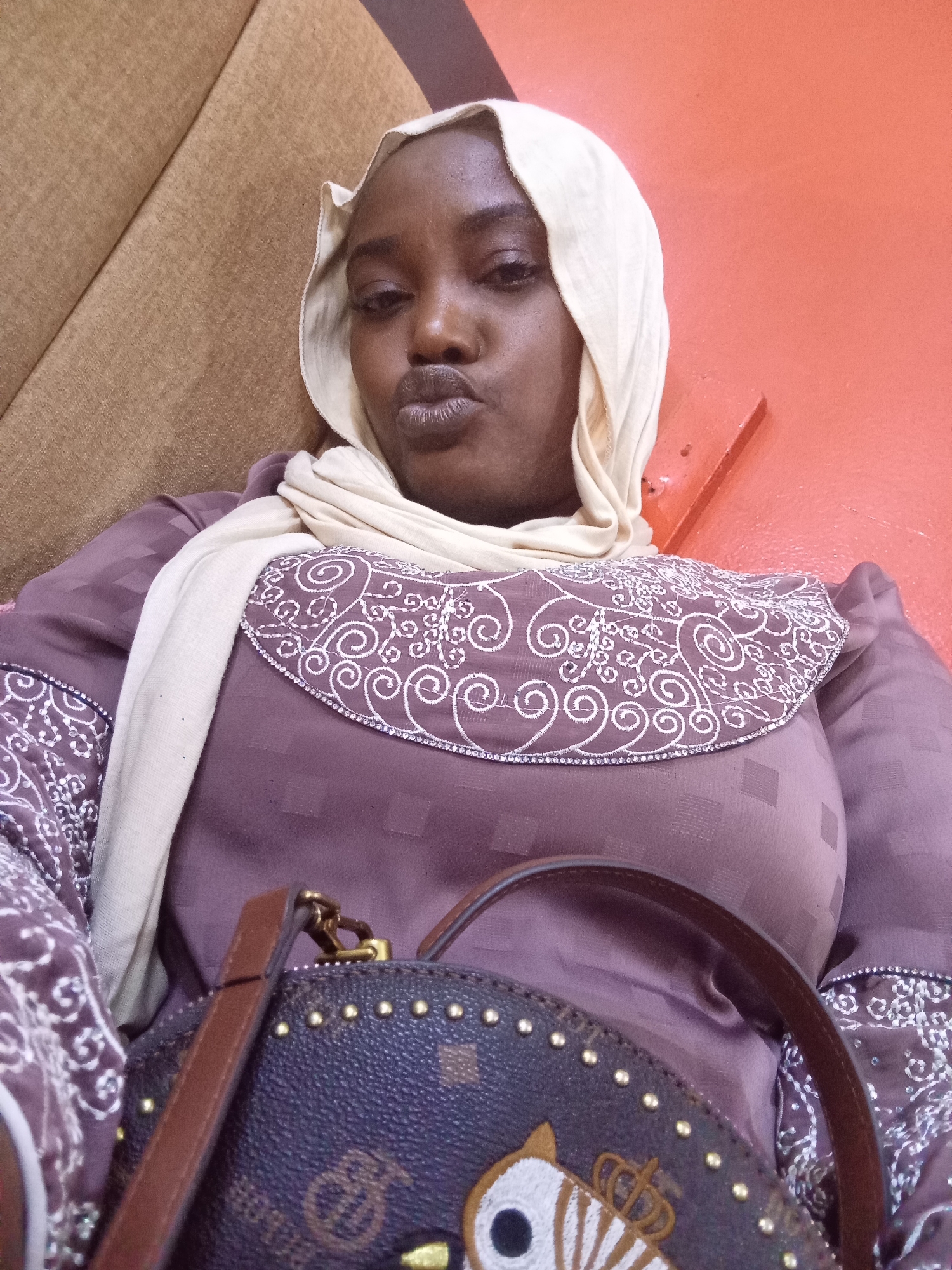A medium shot depicts a woman wearing a light beige headscarf, a mauve dress with white patterned trim, and a dark-colored, studded purse. The woman is seated, leaning against a tan-colored surface with an orange wall visible in the background. Her lips are pursed. The location is in the city of Ajiwa, Nigeria.
