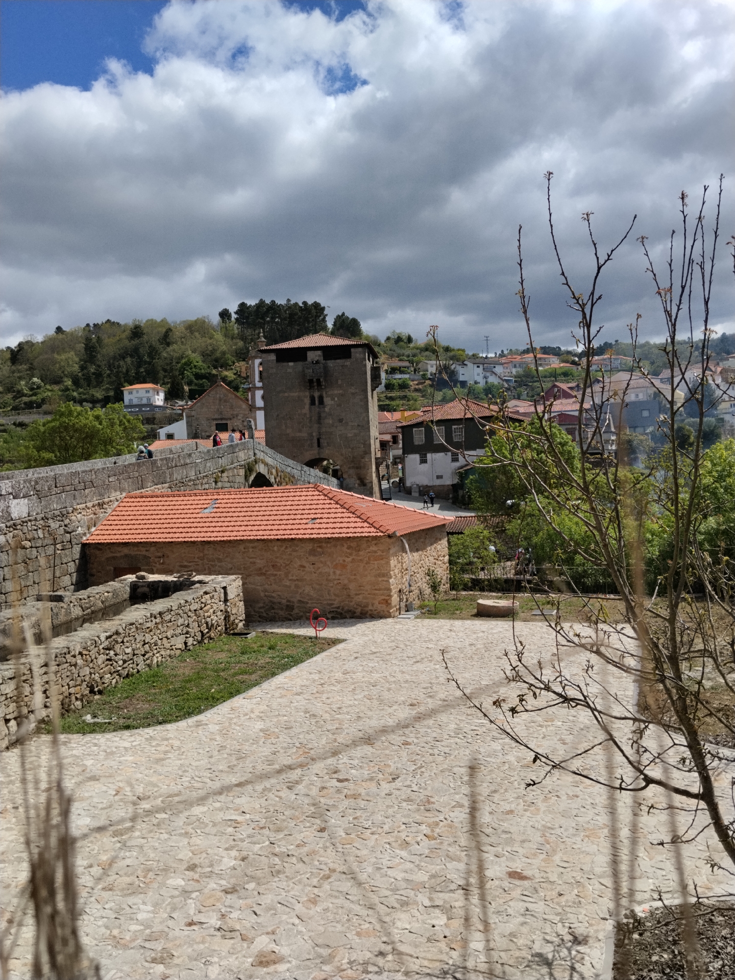 The image depicts a cobblestone plaza in Tarouca, Portugal. A stone building with a red tile roof and a stone aqueduct are visible. In the background is the Ponte Fortificada de Tarouca, a fortified bridge. Several figures are walking across the bridge. Residential buildings are nestled on the hillside in the background. A bare tree with budding leaves is in the foreground, partially obscuring the view. The sky is overcast.