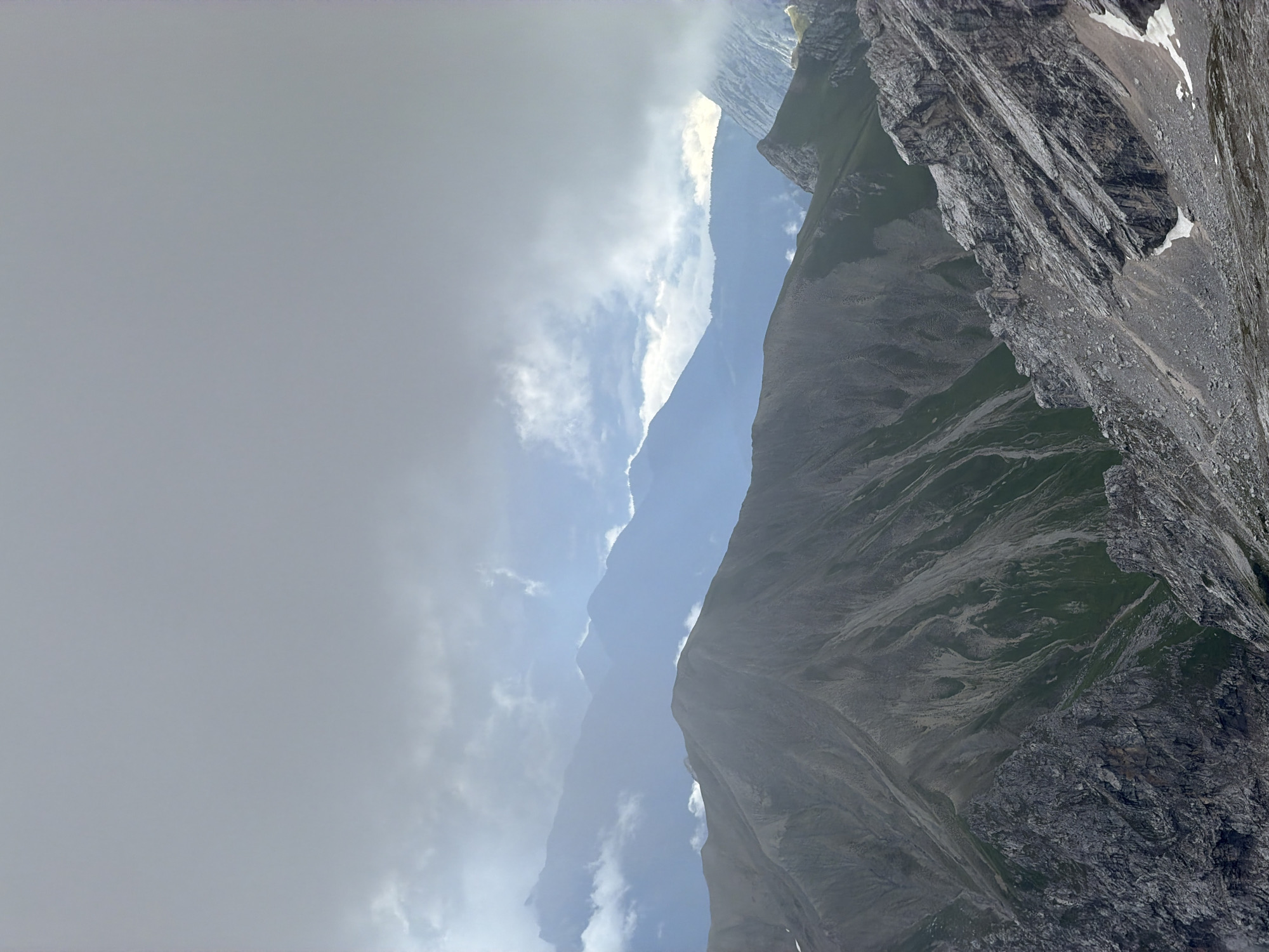 This is a landscape photograph taken from a high vantage point, overlooking a series of mountain ranges under a cloudy sky. The foreground features rugged, rocky terrain with sparse patches of green vegetation, indicative of a steep mountainside. Further back, the slopes are covered in more substantial green grass, transitioning into darker, more muted tones of rock. The background is dominated by layers of mountain peaks fading into a hazy blue, suggesting significant distance. The sky is overcast with heavy grey clouds, but breaks in the clouds reveal lighter, brighter patches, possibly indicating sunlight attempting to break through. The scene is devoid of any people or man-made structures. The overall impression is one of a remote, natural mountain environment, likely captured during the daytime, with atmospheric conditions creating a somewhat somber or dramatic mood.
Based on the visual characteristics and common viewpoints in the region, this scene is consistent with the mountainous landscape found around Garmisch-Partenkirchen, Germany, specifically a view from a higher peak looking towards other mountain ranges within the Bavarian Alps.
