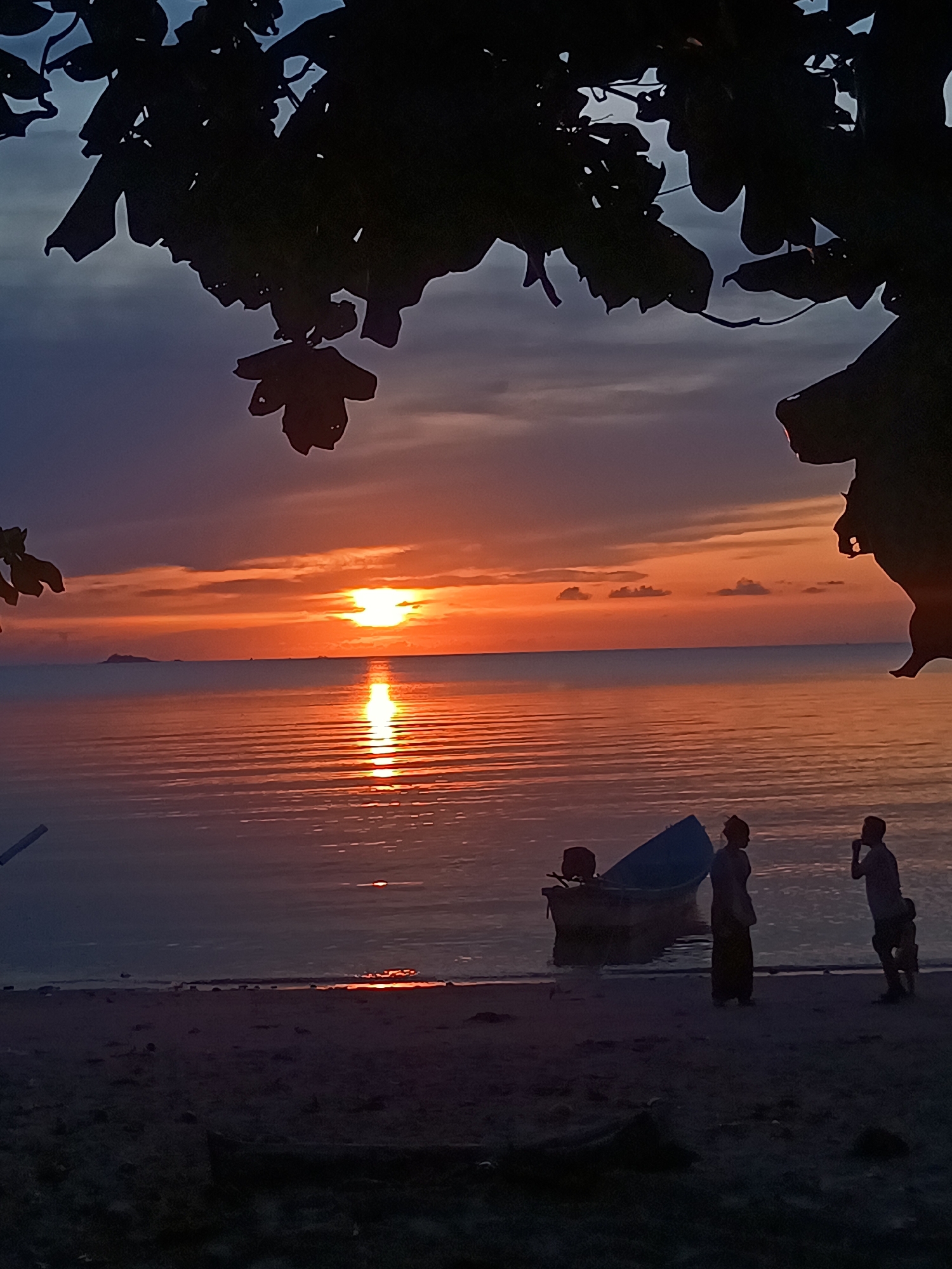 Captured near Baan Wok Tum, Thailand, the image depicts a sunset scene at the beach. The sun reflects off the water, creating a bright line. Silhouettes of three people are visible on the beach, along with a small boat. Above, tree foliage frames the scene.