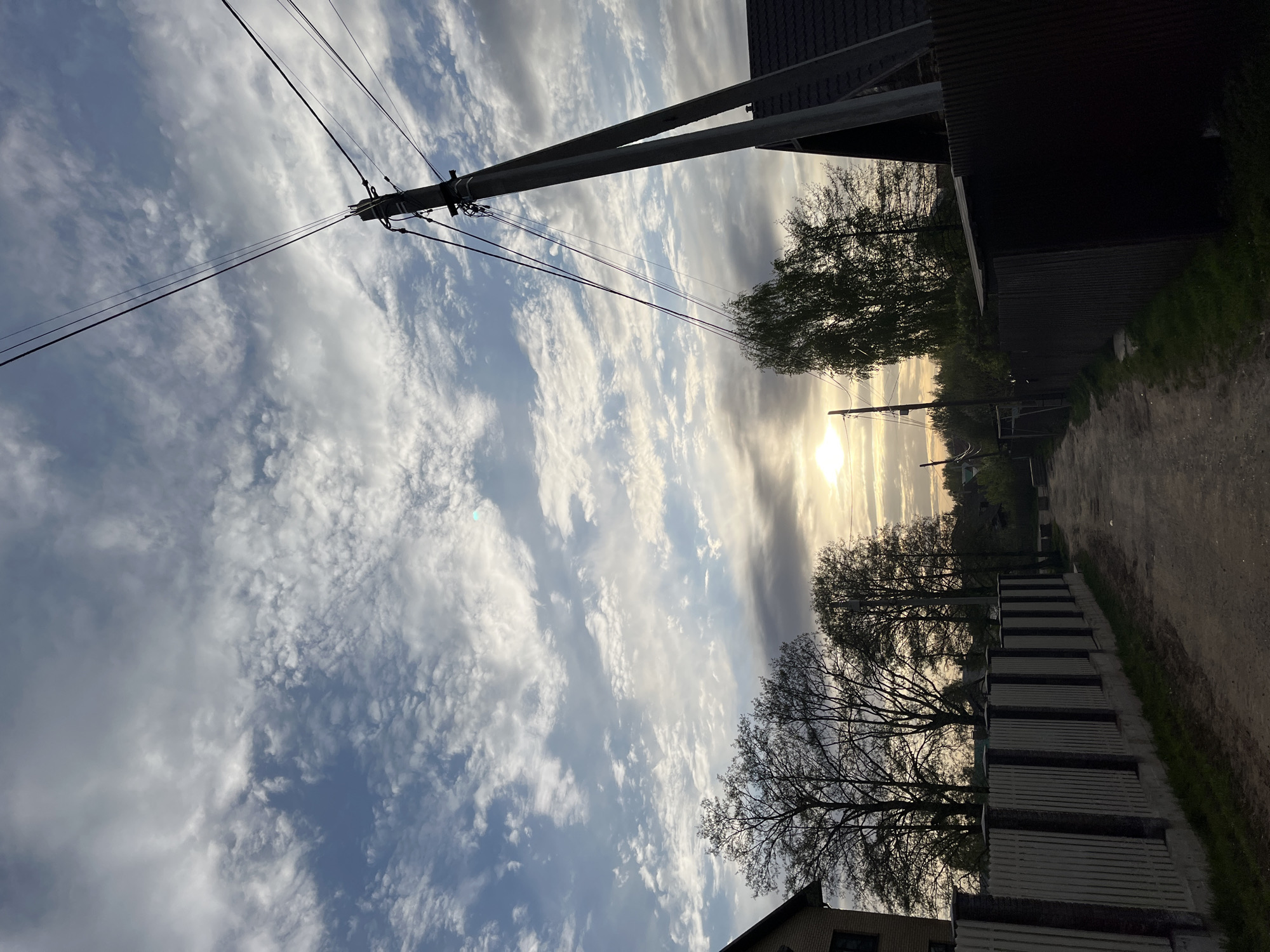 The image shows a street scene in Chaikovsky, Russia at sunset. The view is angled upwards towards the sky. An electrical pole with wires is prominently featured on the left. The sky is filled with streaks of clouds, varying from light, fluffy clouds to dark, dense formations. The sun is positioned in the distance, casting a bright light. On the right side of the image, a dirt road is visible, alongside a fence and trees that line the edge of the road. A building can be seen on the bottom right of the image.