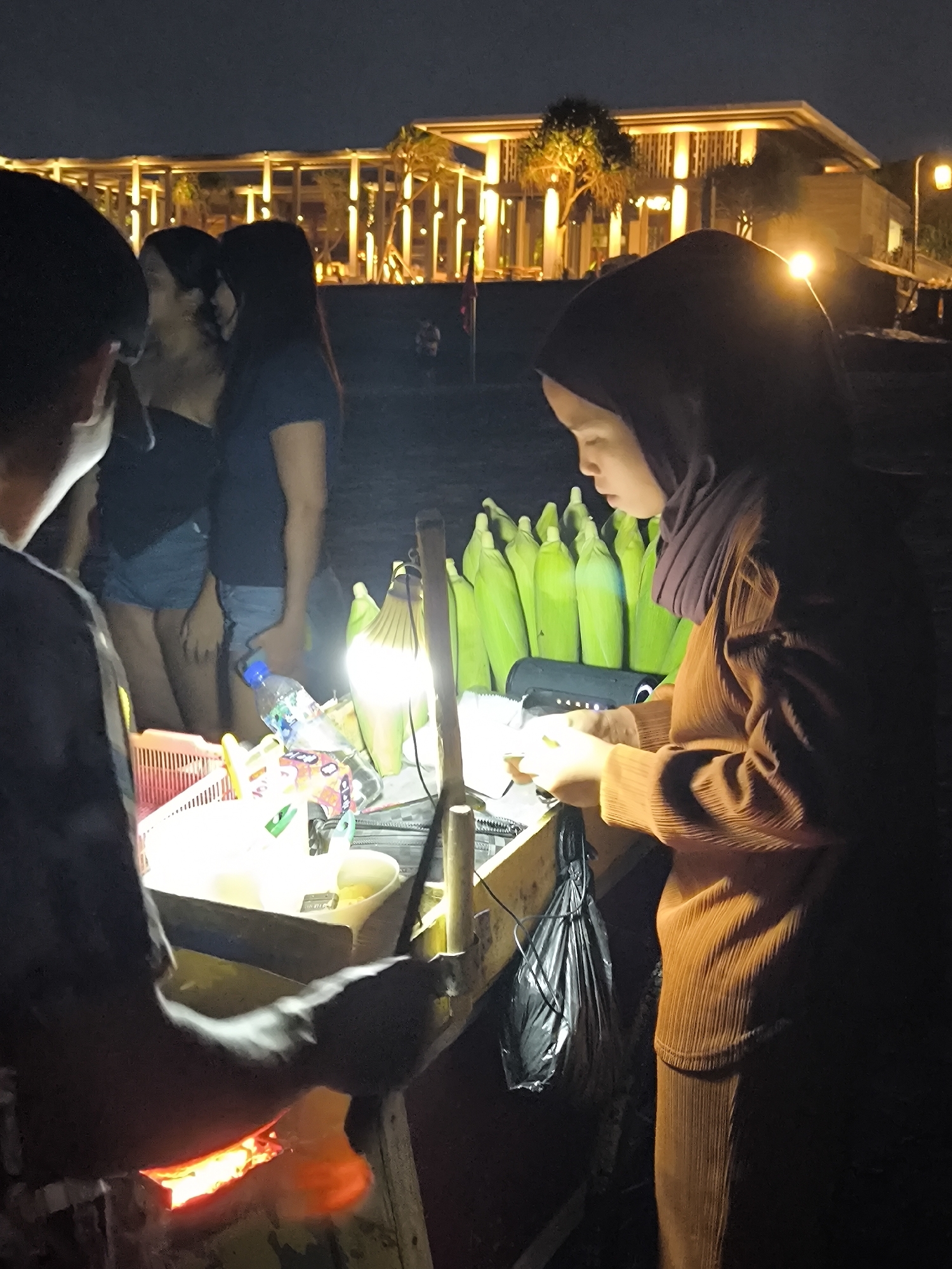 At night in Canggu, Indonesia, a street food vendor wearing a brown hijab and a matching outfit stands behind a mobile food cart. She's preparing corn on the cob, which is stacked on the cart behind her. A light illuminates the cart and the food preparation area. To the left, a person is grilling food on a small grill built into the cart, and a group of people stand nearby. In the background is a modern building with warm lighting and trees. A street light is visible to the right of the building.