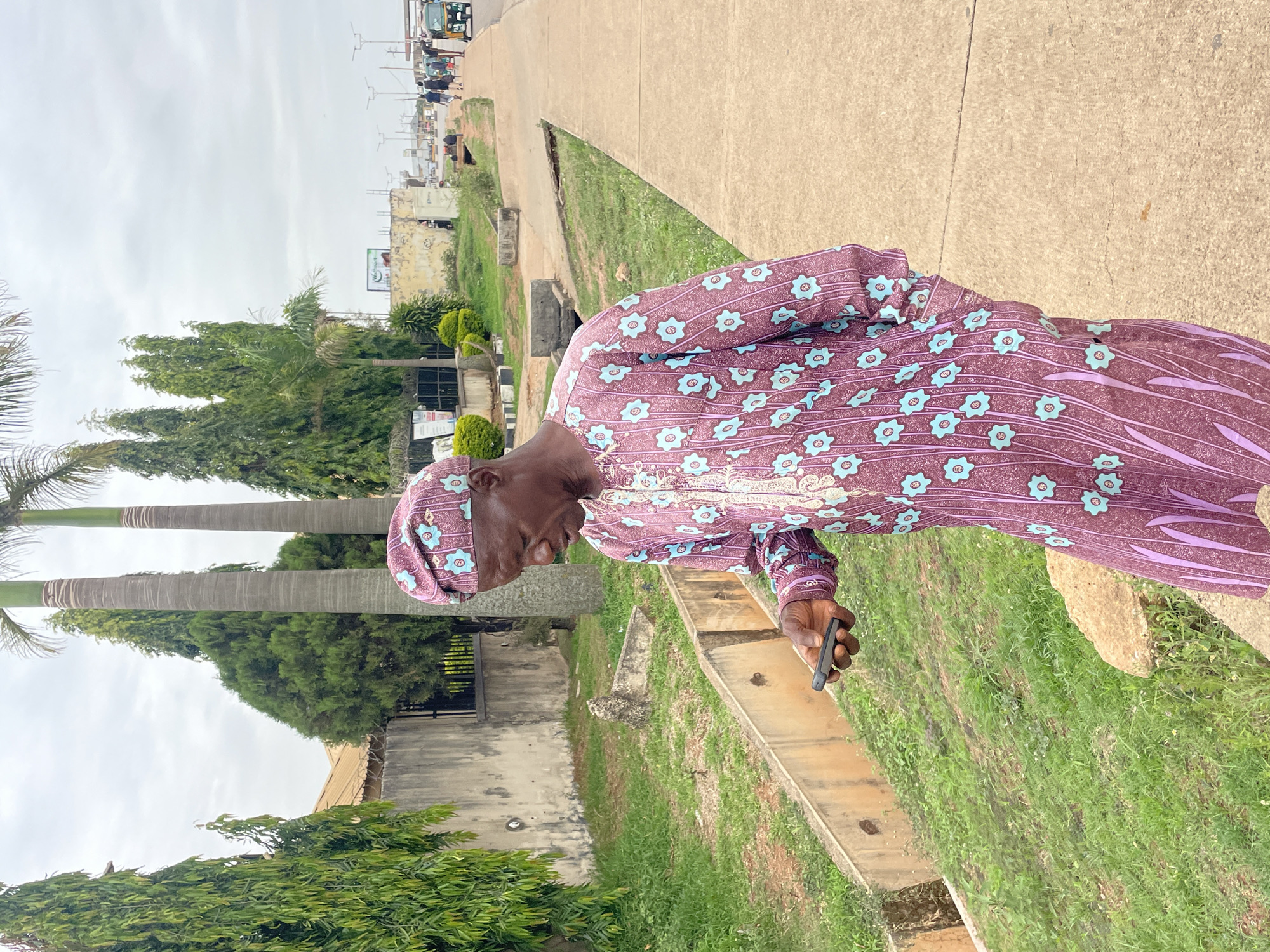 A man in traditional clothing stands on a sidewalk near a road, holding a cell phone. The background includes buildings, trees, and vehicles. The scene is likely in Jos, Nigeria.