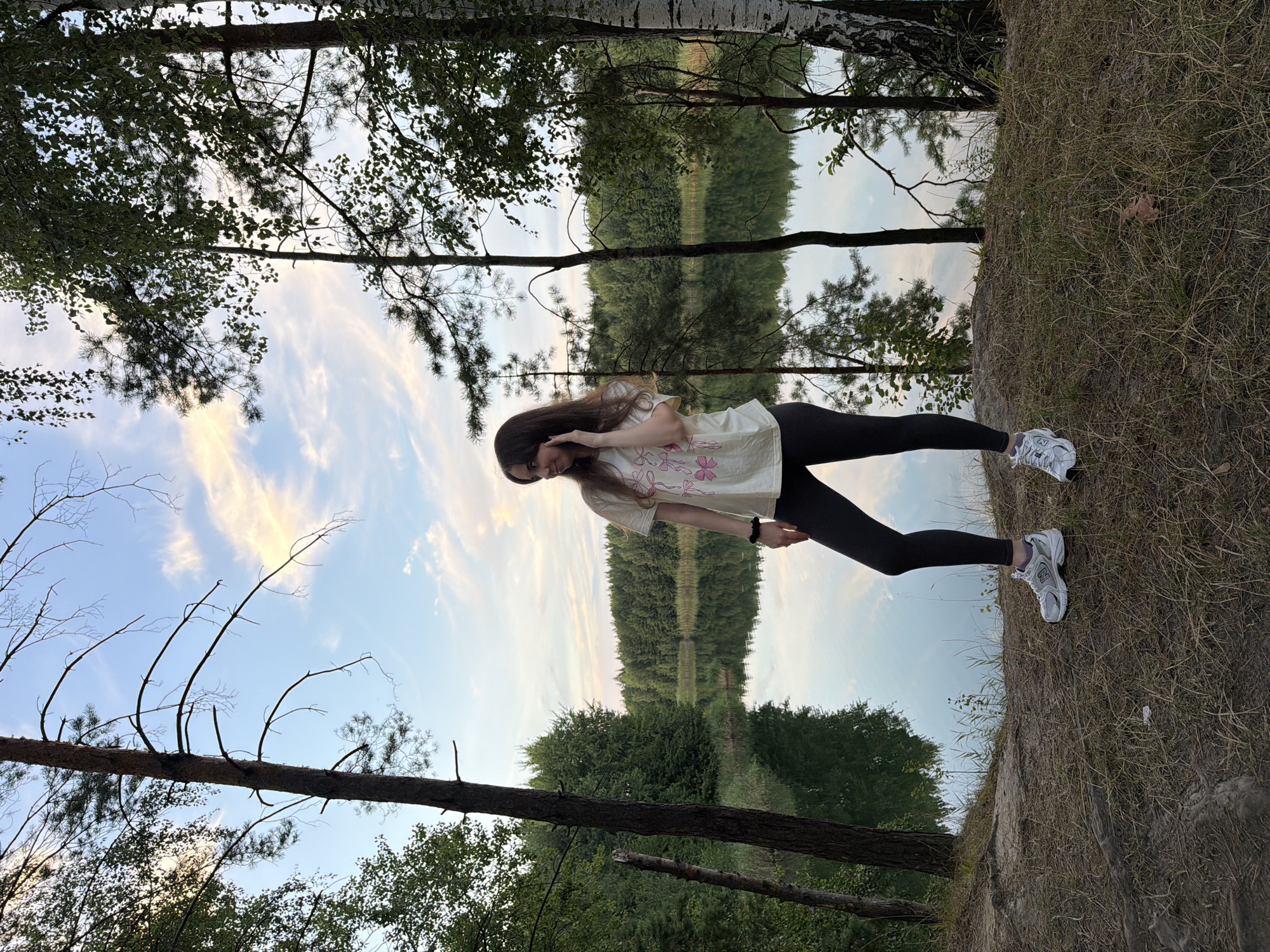 A full-body shot captures a young woman standing near a body of water in Kmytiv, Ukraine. She is positioned on a grassy embankment, facing left and looking towards the water. The woman has long, dark hair and is wearing a light-colored t-shirt with pink bows printed on it, dark leggings, and white athletic shoes. Her right hand is raised to her ear, and her left arm rests by her side.

The background features a serene lake reflecting the sky and surrounding trees. The water is calm, with a visible shoreline lined with dense green foliage. Tall trees, including pines and birches, frame the scene. The sky above is a mix of pale blue and soft pinkish-white clouds, indicating either dawn or dusk. The ground in the foreground is covered with dry grass and sparse vegetation. The overall environment suggests a natural, wooded area.