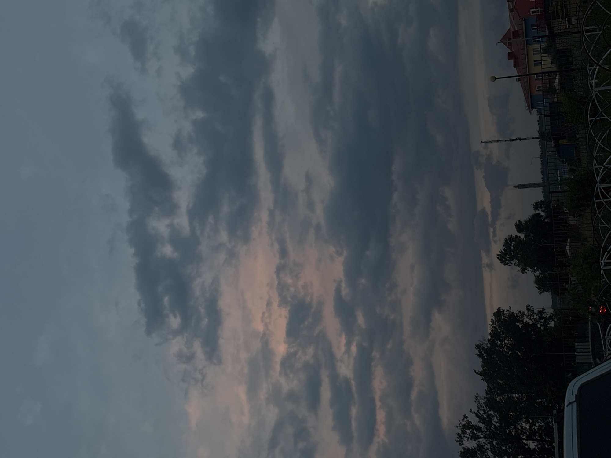This media file shows a cloudy sky over a residential area in Mikhaylovsk, Russia. The sky is filled with dark, streaky clouds that partially obscure a lighter, possibly sunset-tinged sky. In the background, several multi-story buildings with colorful facades are visible, suggesting a residential or possibly a school setting. A street lamp stands near the buildings. Trees and a decorative metal fence are also present, bordering the area. The overall atmosphere is dim, implying it is either early morning or late evening, with overcast weather. There is no visible text in the image. The composition is oriented vertically.
